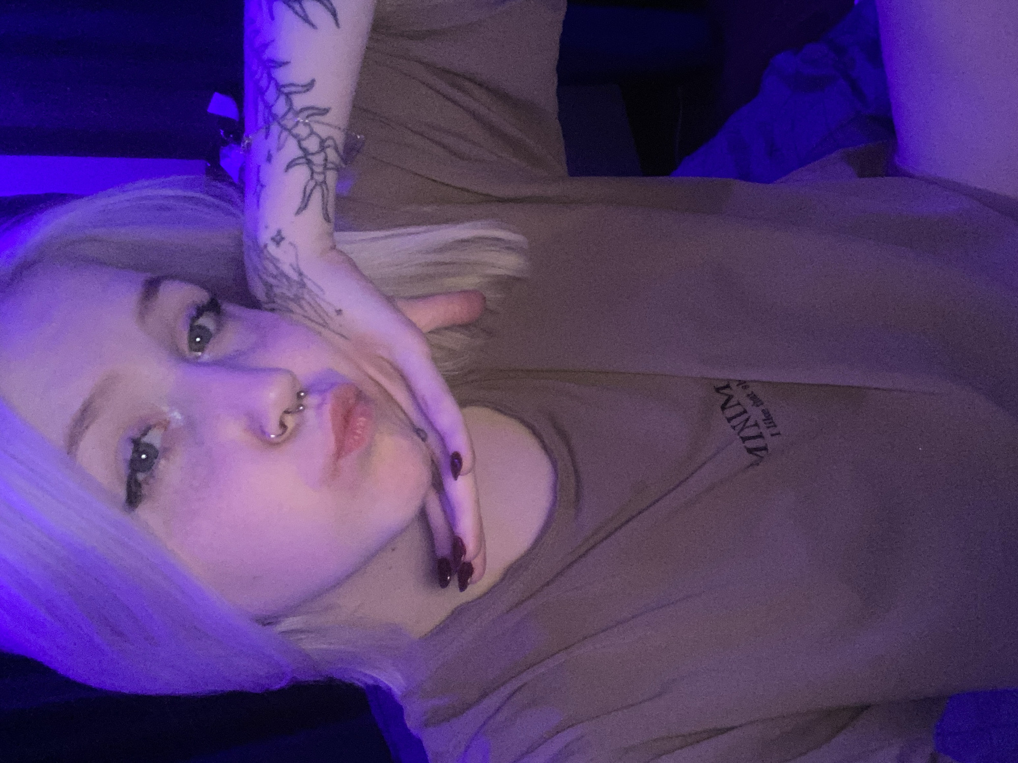 The image shows a woman lying down, with her face tilted towards the camera. She has light blonde hair and a nose piercing. Her right hand is positioned near her face, with burgundy nail polish visible. A tattoo is visible on her right forearm. She is wearing a brown t-shirt with the word "MINIMUM" printed on it. The background includes a mix of blue and purple lighting. There is no location information available in the image or its hints.