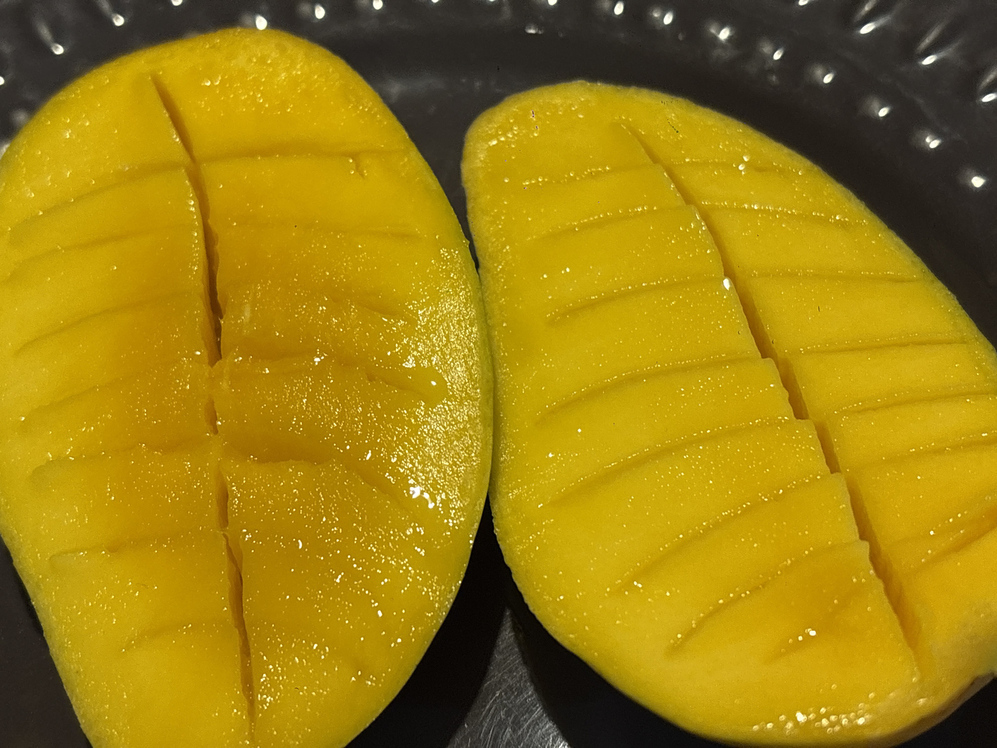 A close-up shot shows two mango halves, each sliced in a grid pattern, placed on a dark, textured surface, potentially a plate. The mangoes are bright yellow and appear ripe. The surface beneath has a dimpled pattern, reflecting light. The location appears to be indoors. The image does not suggest any information regarding Mexico City.