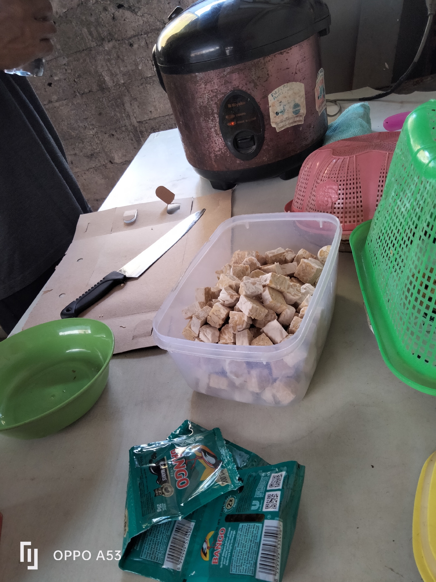 This is a medium shot, eye-level, close-up, indoor photograph of a table with food preparation items. In the background, a distressed, older model rice cooker with a brown and black exterior sits on a counter. To its right are two plastic baskets, one pink and one green, and a light blue towel. In the foreground, a green plastic bowl is on the left side of the table. To its right, a silver knife with a black handle rests on a piece of cardboard. Next to the knife, a clear plastic container is filled with cubed tempeh. In the bottom right, two green packets of "Bango" seasoning are stacked. The table surface is a light beige color, and the background wall is a rough, grey concrete. There are no visible people in the frame. This scene is consistent with a domestic or small-scale food preparation area, likely in Denpasar, Indonesia.