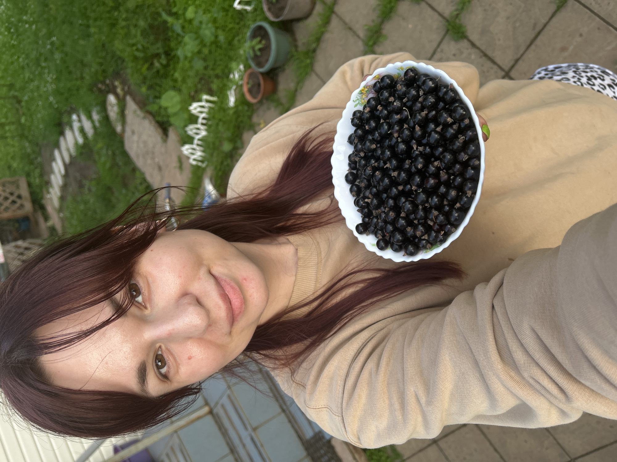 A woman with dark hair, wearing a beige sweatshirt, smiles at the camera while holding a white bowl filled with black currants. Her nails are painted a bright green. The image appears to be taken outdoors, with hints of a garden and paved ground visible in the background. The scene suggests a casual, possibly domestic setting. The lighting implies it is daytime, and the overall mood is cheerful. There is no visible text in the image.