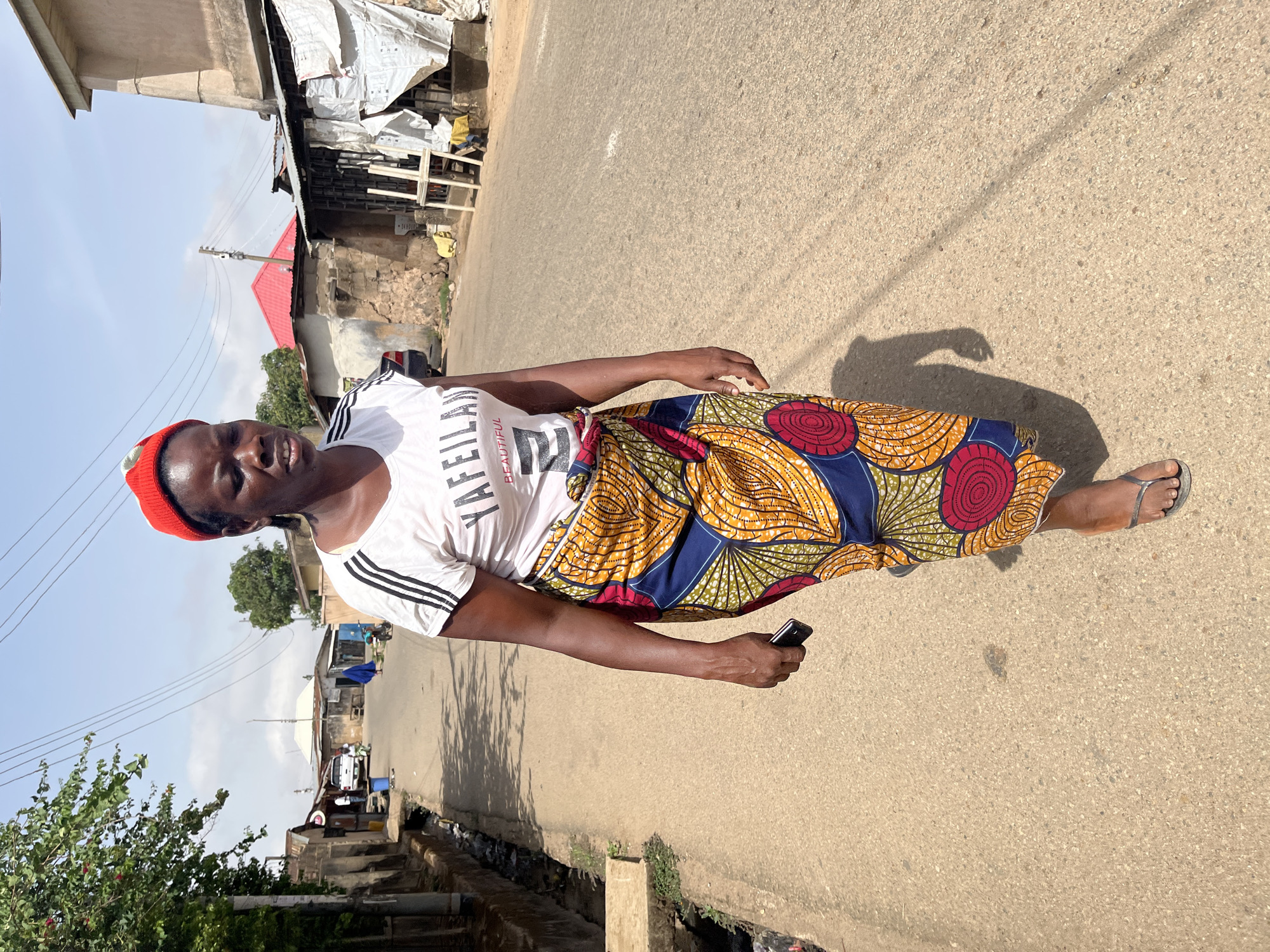 A woman stands on an asphalt road in Jos, Nigeria. She wears a red beanie, a white t-shirt with black stripes on the sleeves, and a long, patterned skirt in yellow, red, and blue. She holds a black smartphone in her right hand. Buildings with corrugated metal roofs and a red roof are in the background, and a few vehicles are visible in the distance. A drainage ditch runs along the side of the road. The sky is pale blue, and the scene appears to be bathed in sunlight.