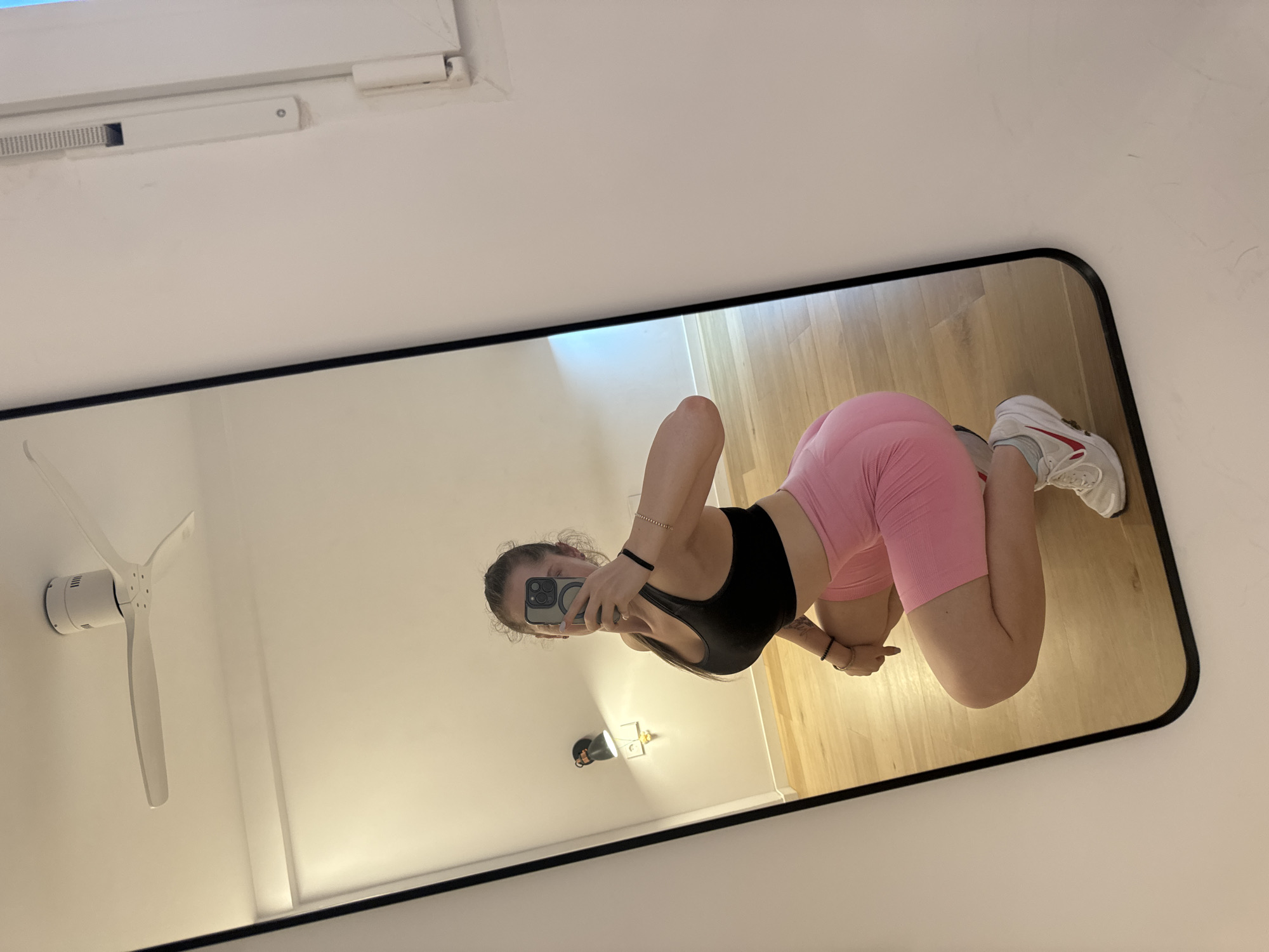 A young woman with brown hair, wearing a black sports bra and pink biker shorts, is reflected in a full-length mirror. She is in a squatting position, holding a smartphone in her right hand to take the selfie. Her left arm is bent, resting on her knee. She is wearing white athletic shoes with red accents.
The reflection shows a room with light-colored walls and a wooden floor. A white ceiling fan is visible in the upper part of the mirror. A black wall-mounted light fixture with a gold accent is also reflected on the wall behind her. The overall environment suggests an indoor setting, possibly a home or gym.
The provided information states the location is Murcia, Spain. There are no specific details in the image that confirm this location, such as landmarks or identifying signs. The scene is otherwise generic for an indoor setting.