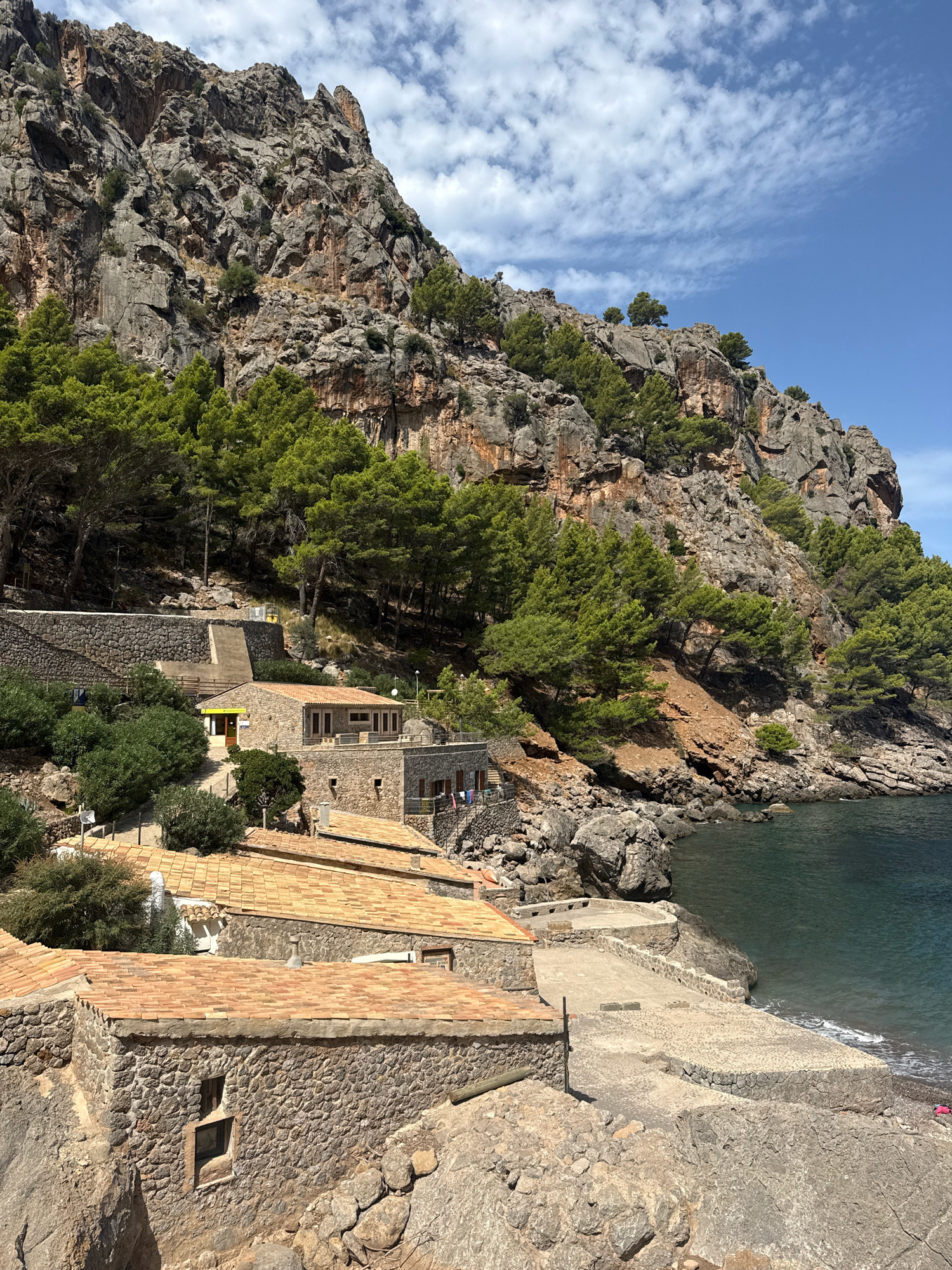 This image captures a scenic view of the coastal area in Port De Sa Calobra, Spain. The foreground features several stone buildings with terracotta tile roofs, built into the rocky hillside. One building in the immediate foreground has a small, recessed window. Adjacent to these structures is a concrete pathway leading down to the water. The sea is a dark blue-green, with gentle waves lapping against a rocky shore and a small pebble beach. In the background, a towering, rugged cliff face dominates the landscape, covered in dense green pine trees. The sky is a clear blue with scattered white clouds.