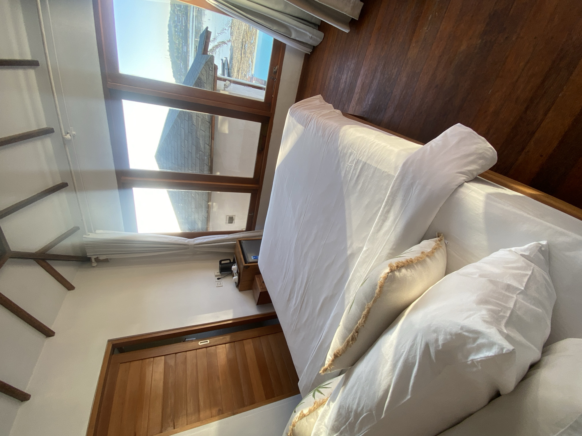 This media file is a first-person perspective photograph of a bedroom with a large window overlooking a body of water and a coastline. The room features a wooden floor, white walls, and a bed with white linens and several pillows. Sunlight streams in through the window, illuminating the room and casting shadows. There is a closed wooden door on the left side of the frame. On a small wooden shelf near the window, there is a black kettle and what appears to be a mini-fridge. The overall impression is one of a serene and peaceful setting. The natural light suggests it is daytime, likely morning or late afternoon due to the warm light. The view outside indicates a coastal location. The image does not contain any discernible text.