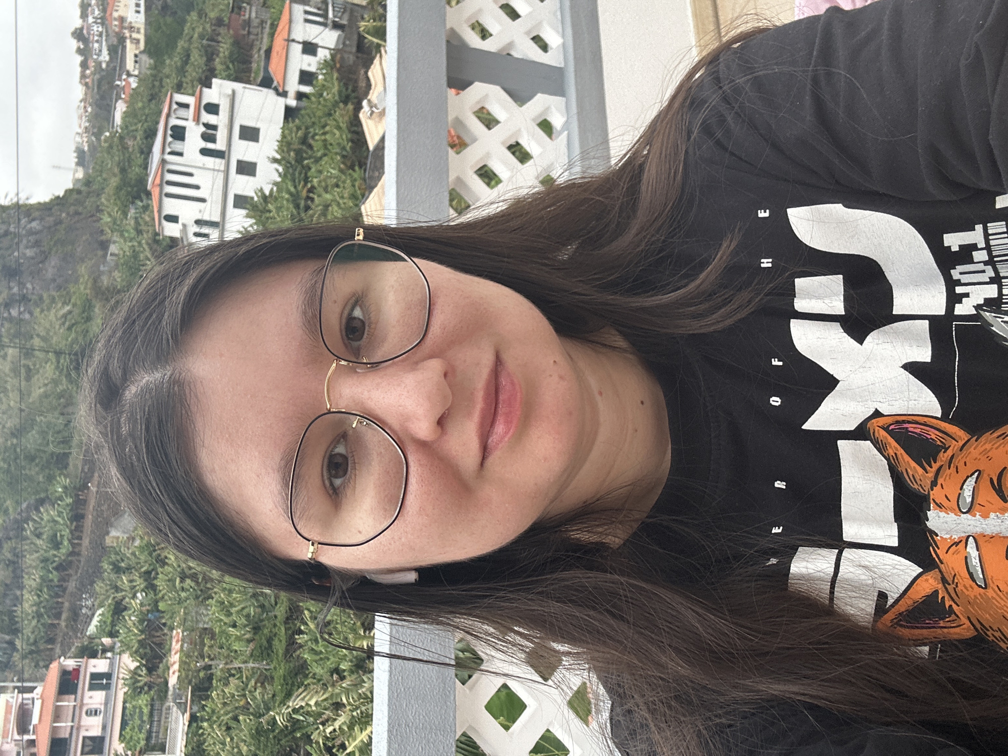 The image shows a person with long brown hair and glasses, wearing a black t-shirt with a white and orange design. The person is positioned outdoors, with buildings and lush greenery visible in the background. The architectural style of the buildings suggests the location is Câmara de Lobos, Portugal. A white lattice railing is visible in the foreground. The overall impression is a casual portrait taken on a balcony or similar outdoor setting.