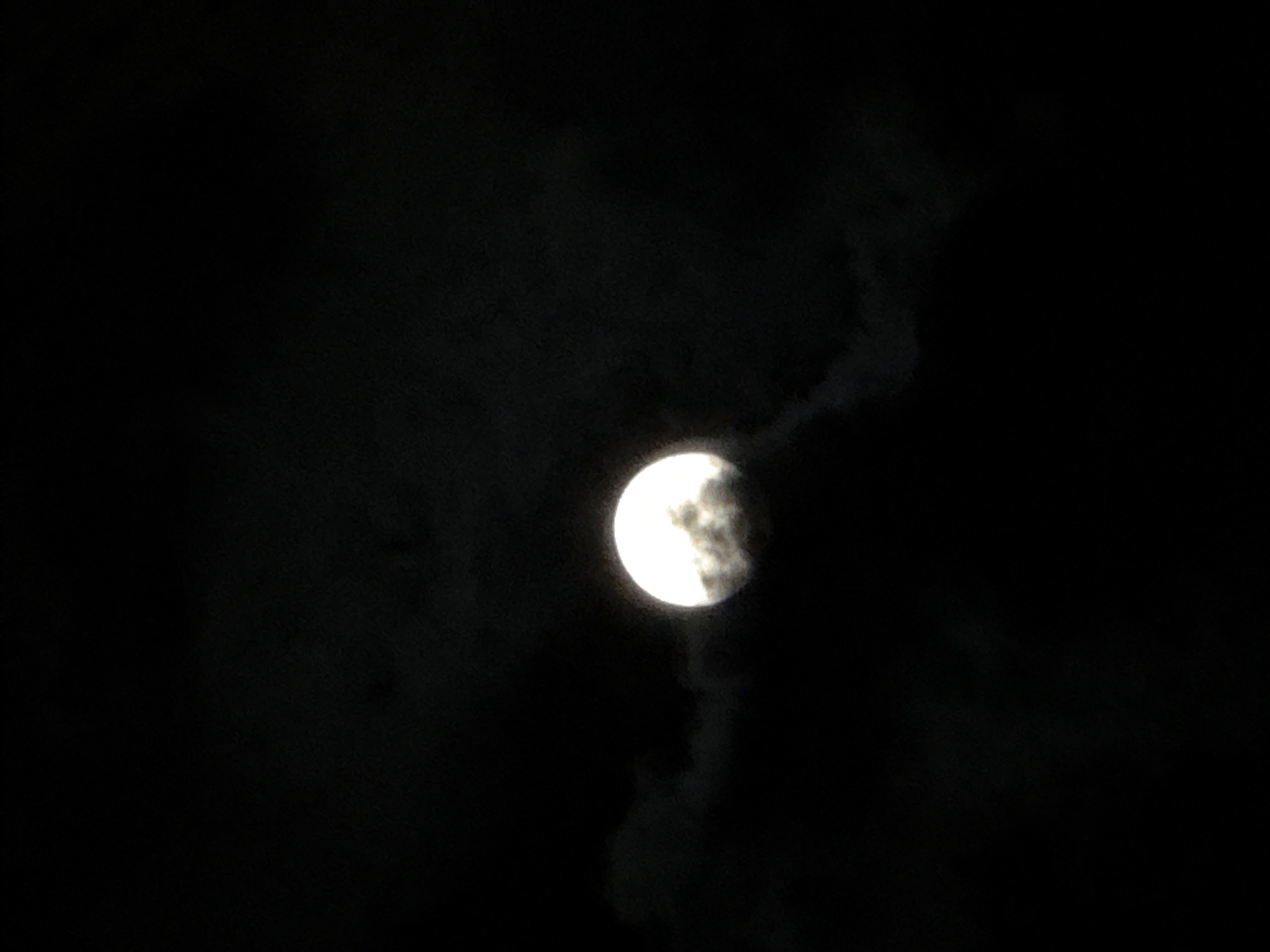 The media file displays a nighttime scene featuring the moon partially obscured by clouds. The main subject is the moon, which appears to be in a gibbous phase, though its exact shape is hard to determine due to the cloud cover. The left side of the moon is brightly illuminated and circular, glowing against the dark sky, while the right side is shrouded by dark, wispy clouds. These clouds have a textured, smoky appearance, extending from the moon's right edge into the surrounding blackness of the sky. The overall setting is a clear night sky with scattered cloud formations, creating a dramatic and somewhat mysterious atmosphere around the moon. There are no visible people, objects other than the moon and clouds, or discernible text in the image. The time of day is clearly night, with the dark sky providing a stark contrast to the luminous moon. No specific location cues are present.