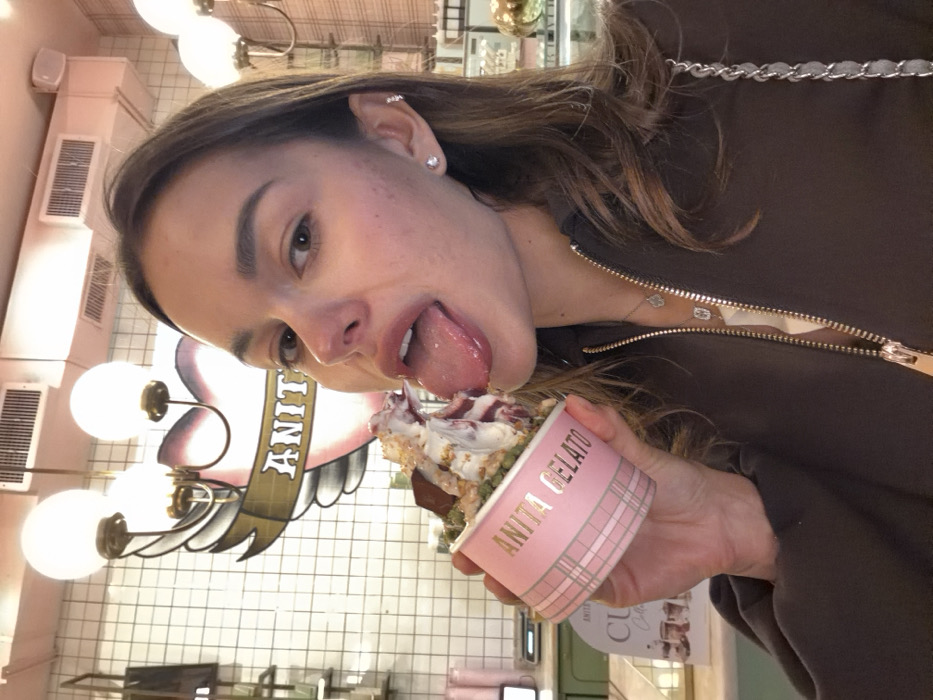 A female individual is depicted in a medium shot, holding a pink "Anita Gelato" cup filled with multiple scoops of gelato and various toppings. Her tongue is extended towards the top of the gelato. She is looking directly at the camera. The individual has long brown hair and is wearing a dark brown zippered top, accessorized with small stud earrings and a silver chain necklace with clover-shaped pendants. The background shows the interior of an Anita Gelato shop, featuring white tiled walls with dark grout, ornate light fixtures with globe bulbs, and a prominent wall logo depicting a winged heart with the name "ANITA" on a banner. A service counter with additional branding is visible to the left. This scene is located within an Anita Gelato establishment in Tel-Aviv, Israel.