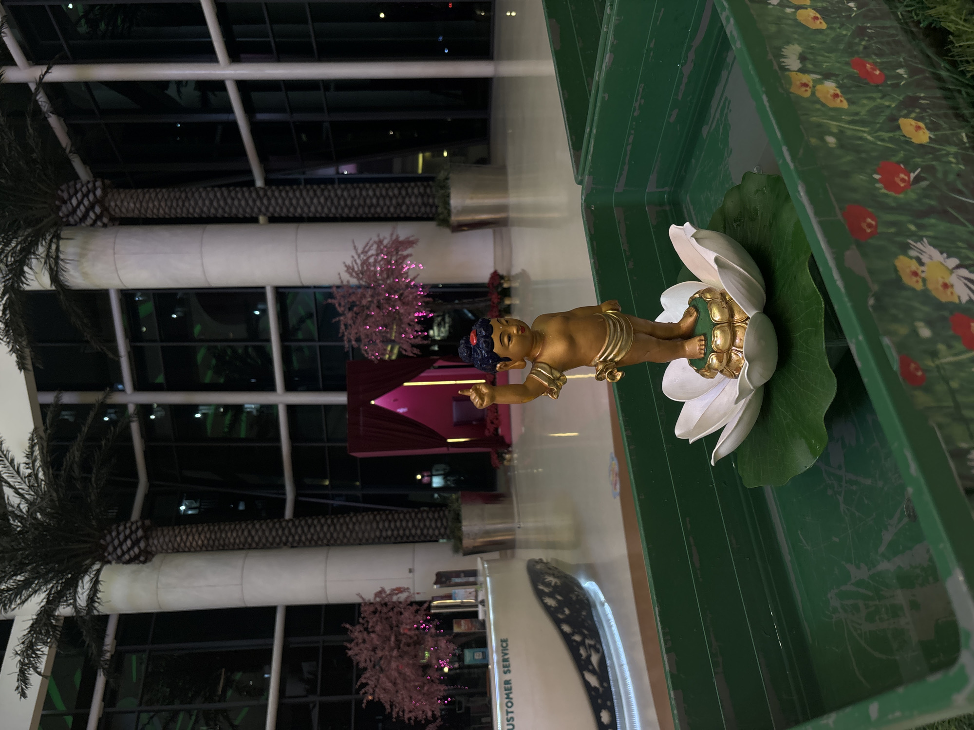 The image shows a golden Buddha statue lying on a white lotus flower inside a green container. The statue is the focal point, with a backdrop featuring artificial trees, indoor building structures, and customer service signage. The scene is located in North Jakarta, Indonesia, as indicated by the cultural context and the overall environment.