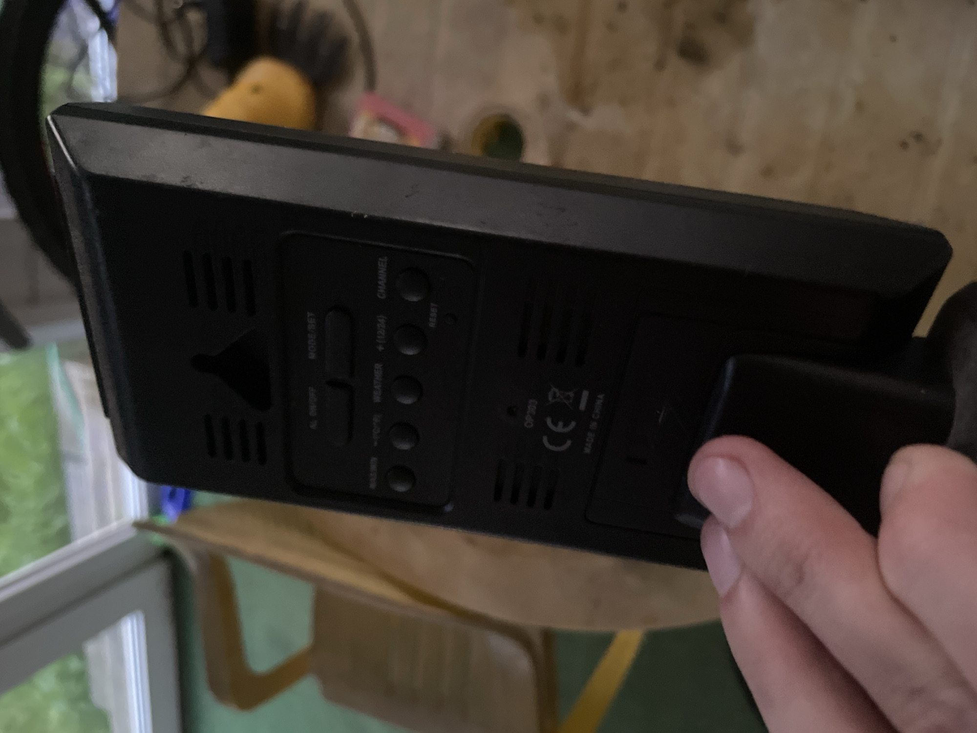 The image shows the back of a black digital weather station. The device has several buttons labeled "AL ON/OFF", "MODE/SET", "MAX/MIN", "-/°F/°C", "WEATHER", "+(12/24)", and "CHANNEL". There is also a small "RESET" button. A "CE" logo and "OP303" are visible, along with "MADE IN CHINA". A finger is visible in the foreground, holding onto a black battery compartment. The weather station is placed on a wooden surface, possibly a table, with a blurred background that includes what appears to be an indoor setting with natural light and some furniture. The city of Bol'shoe Zaborod'e, Russia is not depicted in this image.