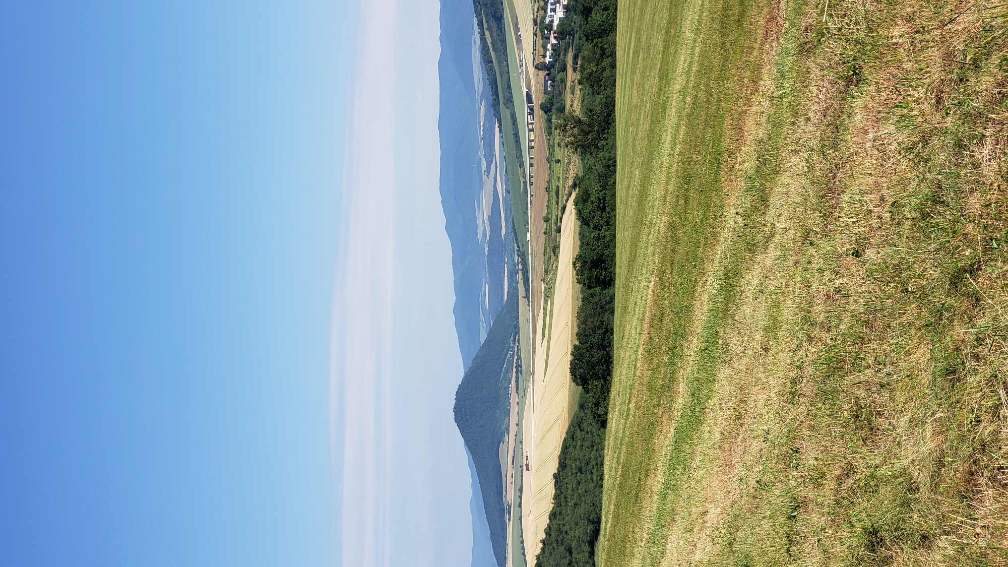 The image depicts a wide, panoramic landscape under a clear blue sky. In the foreground, a mown field of grass, with stripes indicating recent cutting, stretches across the bottom of the frame. Beyond this, a line of dark green trees forms a dense border. Further back, a large, golden field of grain, likely wheat or barley, is visible, with a small building or vehicle appearing as a tiny speck within it.
Dominating the mid-ground is a prominent, cone-shaped hill covered in dark green vegetation. In the distance, a series of rolling hills and mountains recede into a hazy blue. A long, elevated highway bridge with multiple supports spans a valley in the far distance, with some buildings visible near its edge. The overall scene suggests a rural or semi-rural environment, possibly in the vicinity of Prešov, Slovakia, given the geographical features. There are no people or animals visible in the image. The lighting indicates a bright, sunny day.