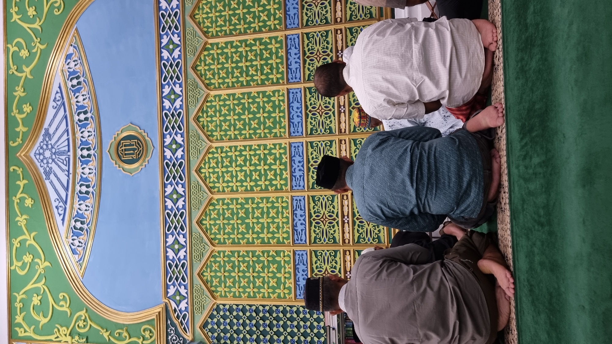 In the city of Keudekruenggeukueh, Indonesia, a group of men are seen from behind, kneeling on a green carpeted floor in prayer. They are facing a wall adorned with intricate Islamic calligraphy and geometric patterns in shades of blue, green, and gold. The men are dressed in traditional attire, including peci hats, and appear to be engaged in a religious service or personal devotion within a mosque or similar place of worship. The wall's design features arched panels with repeating motifs, creating a visually rich backdrop for the prayer.