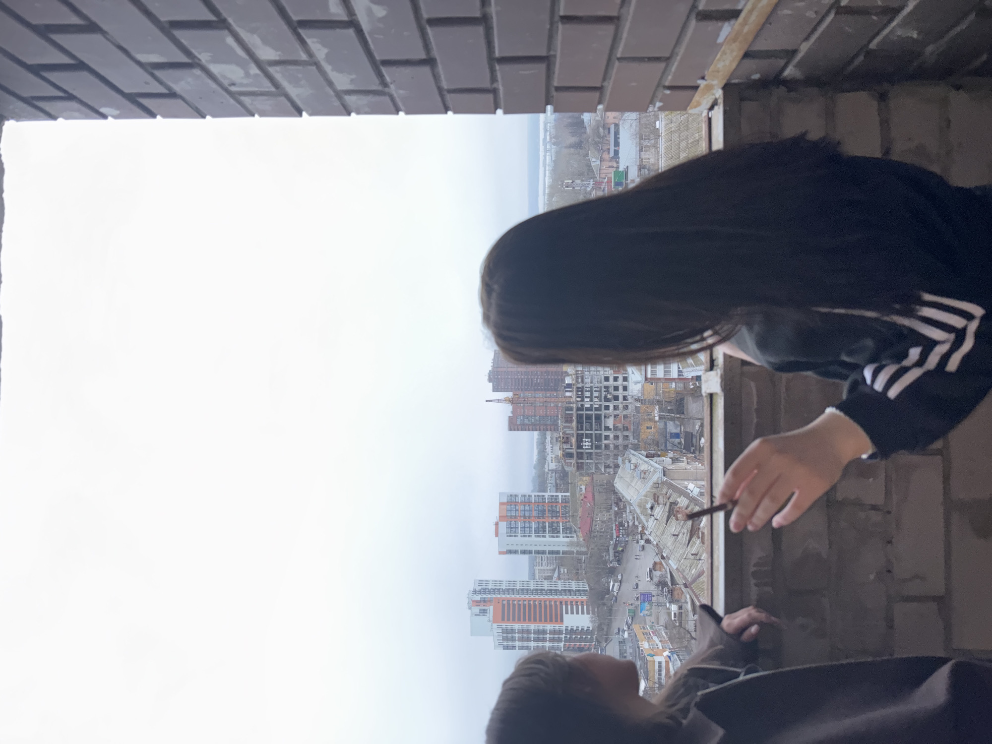 An eye-level shot from an unfinished building in Perm, Russia, capturing two individuals. One person, mostly obscured except for their head and shoulders, is dressed in brown clothing. The other is wearing a black jacket with white stripes on the arms. The visible cityscape features several residential apartment buildings.