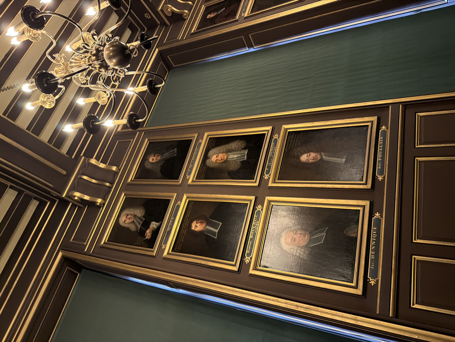 The image displays the interior of a historical institution, likely academic or scientific, located in Paris, France. The perspective is an upward-angled view of a wall and ceiling. The walls feature dark brown wood paneling with intricate gold moldings and decorative elements, interspersed with panels of dark green fabric or wallpaper. A large, ornate multi-arm chandelier, appearing to be brass or bronze and fitted with electric candle-style bulbs, hangs prominently from the equally ornate dark brown and gold patterned ceiling. A significant portion of the wall is covered with a grid arrangement of framed oil portraits. There are at least eight visible portraits, each enclosed in a golden frame, depicting male subjects from the chest up. The men are generally depicted in 17th-18th century attire, including powdered wigs and dark academic or clerical robes with white bands. Visible inscriptions beneath some portraits identify individuals such as "Simon BOULDUC 1729", "Jac. HENNIQUE", and "Ant. POULLAIN". No individuals are actively present or interacting within the depicted scene. The overall lighting is warm, originating from the chandelier.