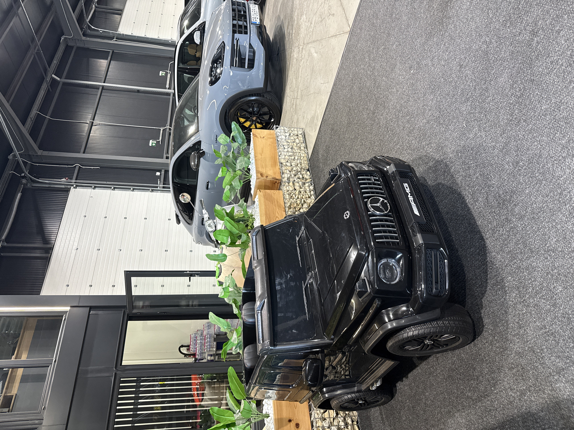 The image depicts a showroom with a black toy Mercedes-Benz G-Class in the foreground and a grey Porsche Cayenne in the background. The toy car is positioned on a dark grey carpet, with its front facing the viewer. The Porsche is on a concrete floor, to the right of the toy car. The showroom appears to be an indoor space with large garage doors in the background and a modern facade with large windows on the left. There are several potted plants and decorative stone elements around the toy car. The image was taken in Plovdiv, Bulgaria.