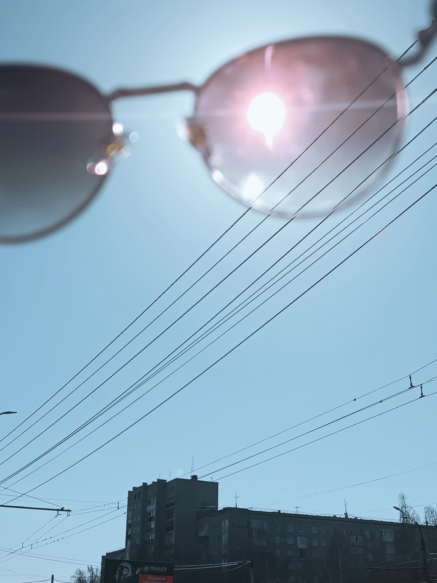 The image depicts a low-angle view looking upward in Izhevsk, Russia. Sunglasses are positioned close to the lens, with the right lens reflecting the sun as a bright, white glare with a pinkish hue. A series of parallel power lines stretch across the sky, converging towards the right side of the frame. Below the power lines, a gray, multi-story residential building is visible. A billboard with the word "МЕДИЦЯ" (MEDITSYA) in Cyrillic and an image of a person is partially visible in the lower left corner. The sky is a clear, light blue color.