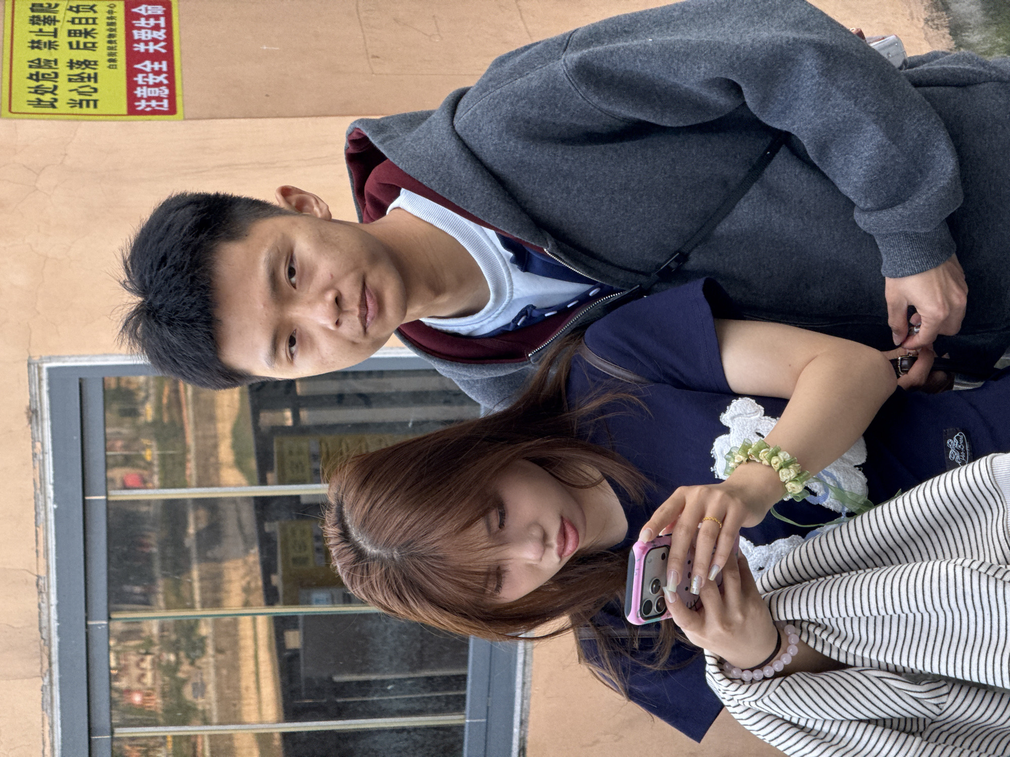 A photograph taken outdoors in Yuzhong District, Chongqing, China shows an Asian man and woman sitting, facing the camera. The man on the left has short black hair, tan skin, and is wearing a grey zip-up hoodie with a maroon lining, a white t-shirt, and a blue shirt underneath. He is looking directly at the camera. The woman on the right has shoulder-length brown hair and is wearing a dark blue t-shirt with a white cartoon image on it, a flower bracelet, and a pink beaded bracelet. She is holding a pink smartphone and looking down at it. A striped black and white shirt lies in her lap. Behind them is a building with a window and a sign in Chinese characters reading "此处危险 禁止攀爬 当心坠落 后里负货 白象街民食物业服务中心 注意安全 关爱生命".