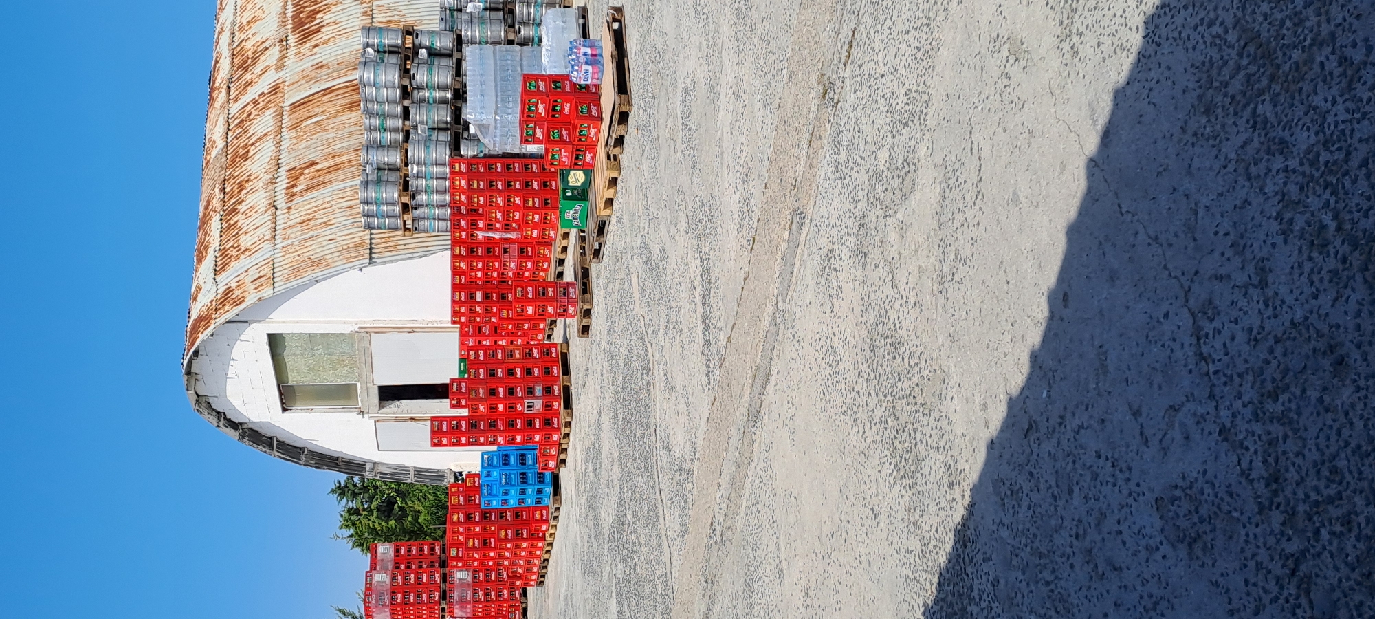 The image depicts an outdoor storage area adjacent to a weathered, arched-roof building in Velika, Bulgaria. The building, with its corrugated, rusted roof and white facade, appears to be a warehouse or storage facility. In front of the building, numerous stacks of red and blue plastic crates are arranged on wooden pallets, predominantly containing beverage bottles. To the right of the crates, several rows of metal kegs are also stacked on pallets, suggesting a brewery or distribution center. The ground is paved with a rough, textured surface, and a long shadow stretches across it from the left side of the frame, indicating the presence of an unseen object or structure. The sky is clear and blue. No people are visible in the image.