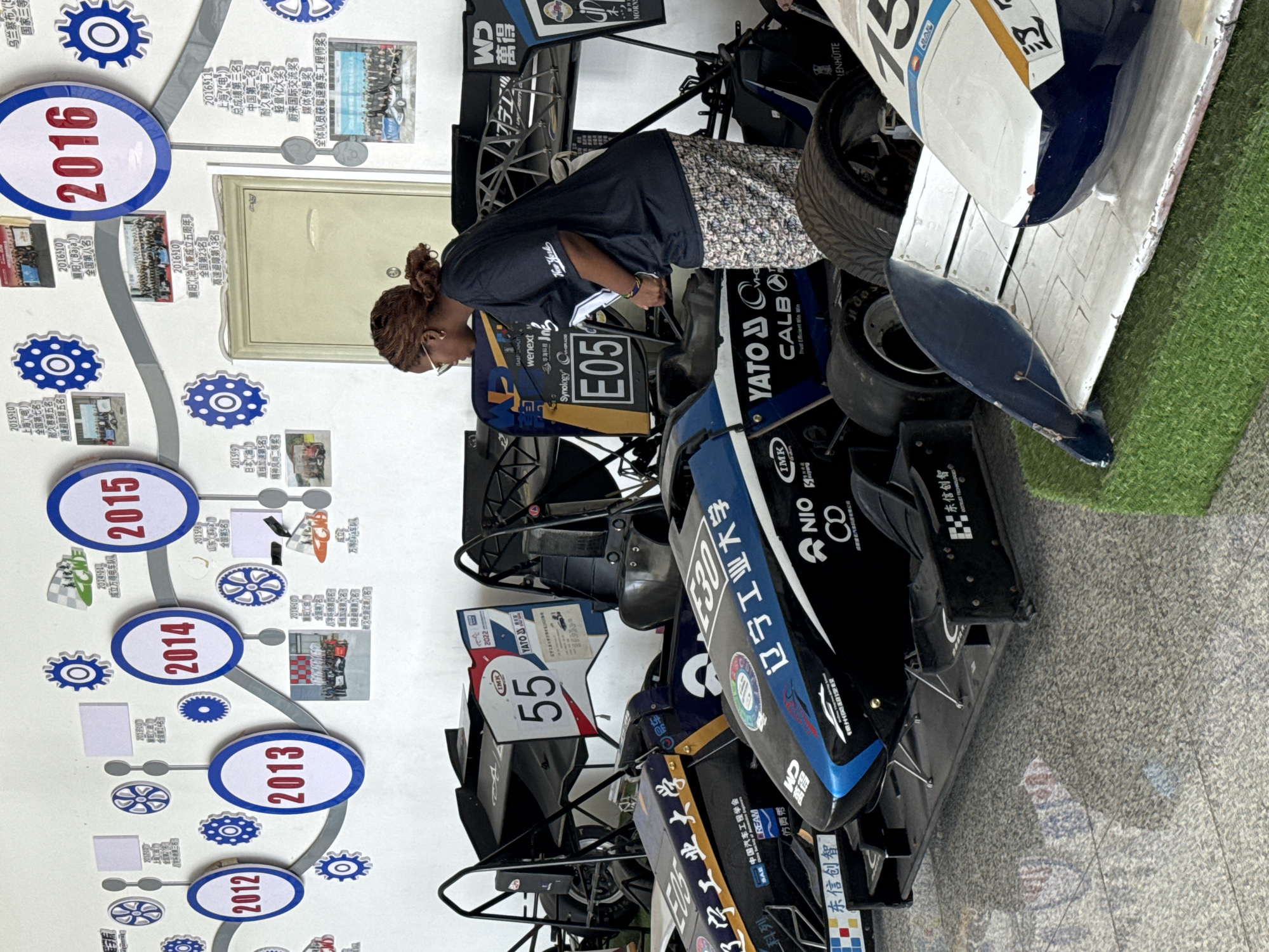 This is an eye-level medium shot of a young Black woman wearing glasses, a dark t-shirt with white lettering, and patterned capri pants, standing in front of three racing cars and a timeline display on a wall. The woman is looking down and to her right, appearing to examine the closest racing car. Her hair is styled in a bun. The racing cars are positioned closely together. The closest car is predominantly black with blue and yellow accents and features several logos and text in Chinese, including "辽宁工业大学" (Liaoning University of Technology). A number "E05" is visible on the side of this car. The car behind it is black and blue with similar Chinese lettering. The third car is white with blue accents and a red and white checkered flag design on its rear wing, with the number "15" visible. The timeline on the wall behind the cars is a historical display with circular markers for years such as "2012", "2013", "2014", "2015", and "2016", accompanied by small photographs and text in Chinese. The environment appears to be indoors, possibly a workshop or exhibition space, with a light-colored wall and a gray, textured floor. The presence of the racing cars and the timeline suggests a connection to an academic institution or racing team. The text in Chinese characters indicates the likely location is in China, and specifically the "Liaoning University of Technology" branding on the cars points to Liaoning Province. Without further context or external information, pinpointing the exact city of Guta within Liaoning Province based solely on the image is not possible.