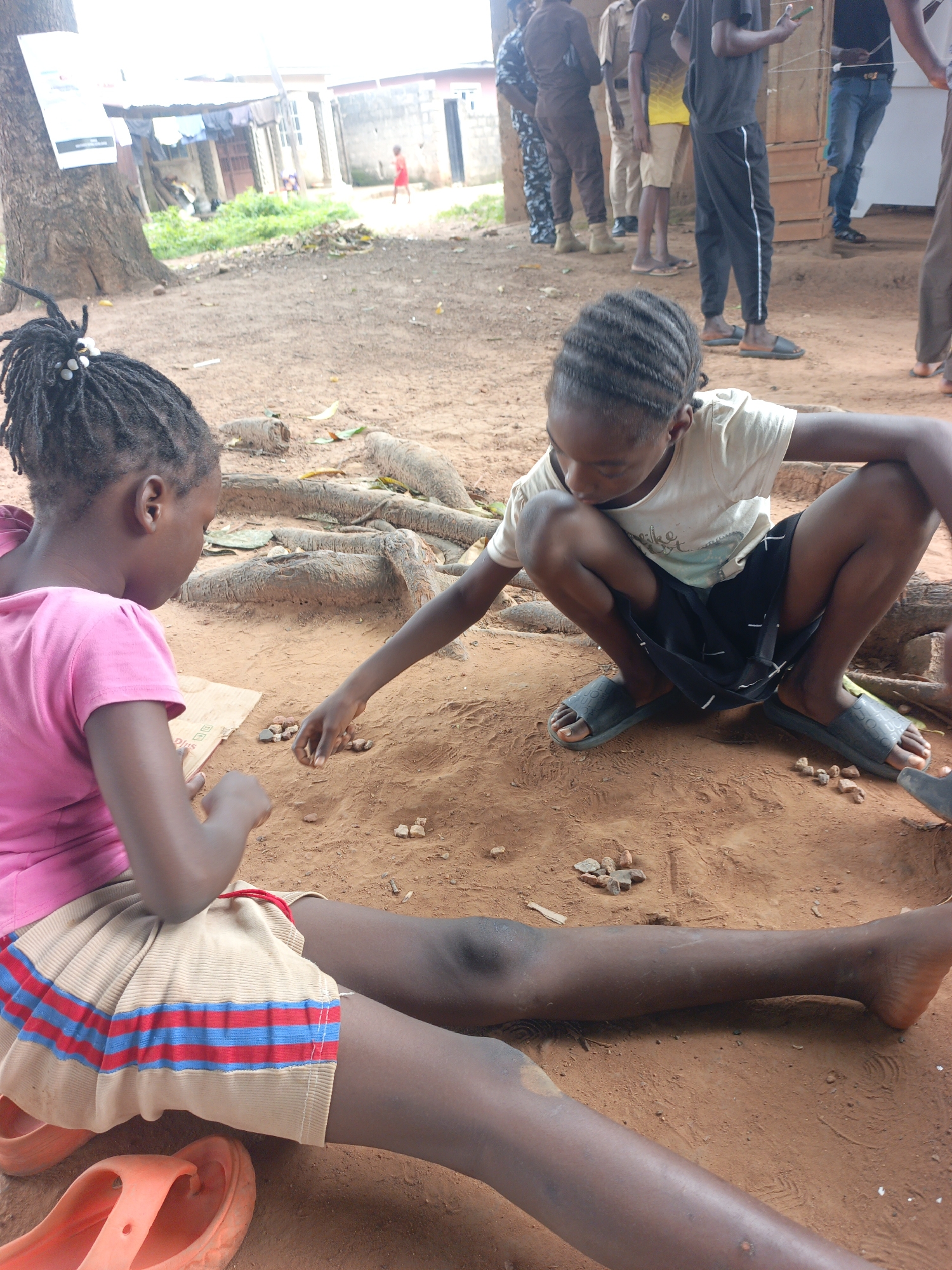 The image depicts two young girls playing a game with small stones on a dirt ground. The girl on the left, wearing a pink t-shirt and a beige skirt with red and blue stripes, is focusing intently on the game. The girl on the right, dressed in a white t-shirt and a dark skirt, is also engaged in the activity. Both girls are barefoot or wearing sandals. The ground is reddish-brown dirt, with exposed tree roots visible. In the background, several adults are standing, some in casual attire and others in what appears to be uniforms. One adult is holding a mobile phone. Behind the adults, there are simple structures, possibly homes or shops, with corrugated metal roofs. Laundry is hanging to dry on a line. The overall setting suggests an outdoor environment in a village or rural area. The lighting indicates it is daytime. The activity appears to be a casual game, and the children's expressions suggest concentration rather than strong emotion. There is some text visible on a poster attached to a tree on the left side of the image, but it is too blurry to read.