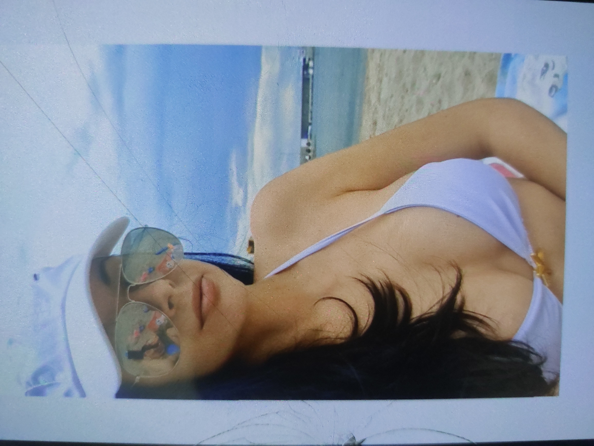 A woman lies on a beach, likely in Tsarevo, Bulgaria, wearing a white bikini and sunglasses. She has dark hair and is wearing a white cap. The background shows a beach with light-colored sand, the sea, and a pier or similar structure. The sky is visible in the upper left corner of the frame.