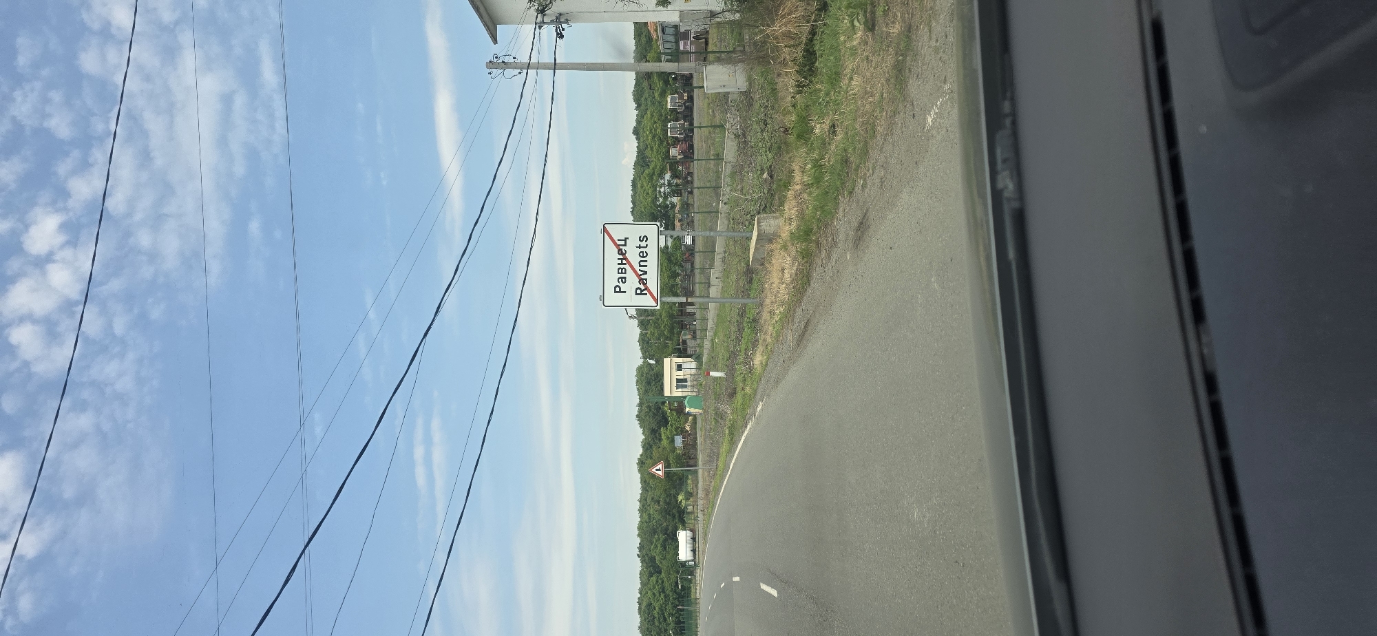 The image depicts an outward-facing view from a vehicle on a paved road, indicating the departure from the settlement of Ravnets, Bulgaria. A white rectangular sign with a red border, prominently displaying "Равнец" in Cyrillic and "Ravnets" in Latin script, is situated on the right side of the road, featuring a red diagonal line indicating the end of the town.
The asphalt road curves gently to the left in the foreground, with a dashed white line marking the center and a solid white line on the right edge. The vehicle's dark dashboard is visible at the bottom of the frame. Overhead, a light blue sky with scattered white clouds is traversed by several black power lines running horizontally across the upper portion of the image.
To the left of the road, a dense line of green trees forms the distant horizon. Closer to the road, a triangular warning sign with a red border and a black exclamation mark on a white background is visible. Adjacent to it, a small, light-colored building with a sloped roof stands. Further left, a white trailer or modular unit is parked.
To the right of the road sign, a wooden utility pole with multiple cross-arms and insulators supports electrical wires. Behind a fence, a collection of various agricultural vehicles, primarily older model tractors, are parked in a row. A portion of a white building with a dark roof is visible on the far right edge of the frame, partially obscured by the utility pole. The roadside is covered with dry grass and sparse vegetation. No individuals are present in the scene.