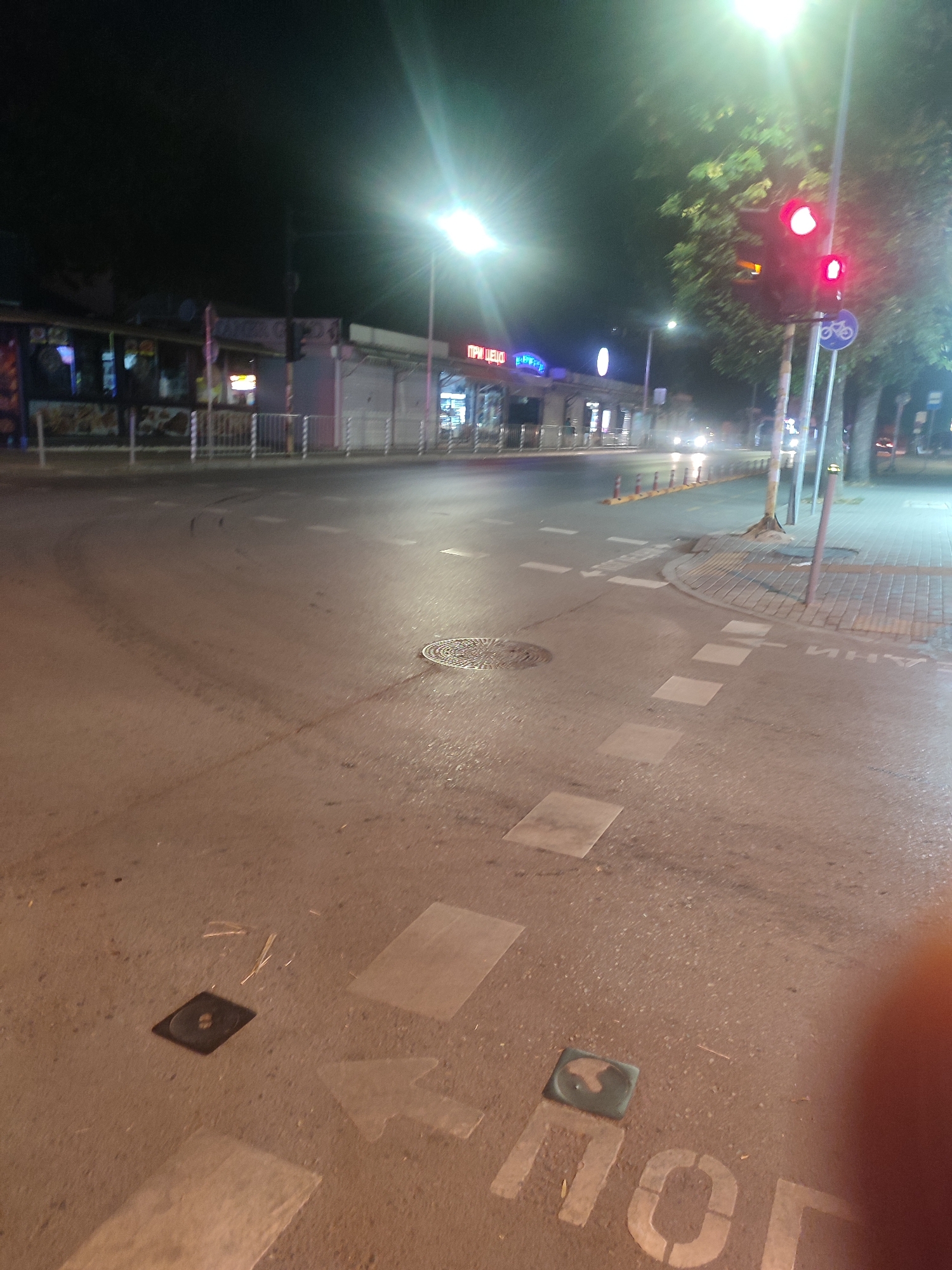 The image captures a street scene at night in Varna, Bulgaria. The perspective is low, looking towards a street intersection with shops and restaurants lining the left side of the road. The shops have bright, neon signage, with one sign clearly visible reading "ПРИ ЦЕЦО" (Pri Tsetso). Traffic lights are visible and illuminated with red lights, indicating a stop. A bicycle lane symbol is present on a blue sign near the traffic lights. The road surface is asphalt, appearing wet, with visible tire marks and road markings, including white painted arrows and squares. There are also manhole covers on the road. In the foreground, white painted letters on the road are partially visible, spelling out "ПОГ" (Pog). The environment suggests a commercial area within the city, possibly a boulevard or a main street with pedestrian access. There are no people prominently visible in the frame. The lighting is a mix of streetlights, shop lights, and vehicle headlights, creating a somewhat hazy atmosphere typical of nighttime urban environments. Trees are present on the right side of the image, their leaves illuminated by the artificial lights.