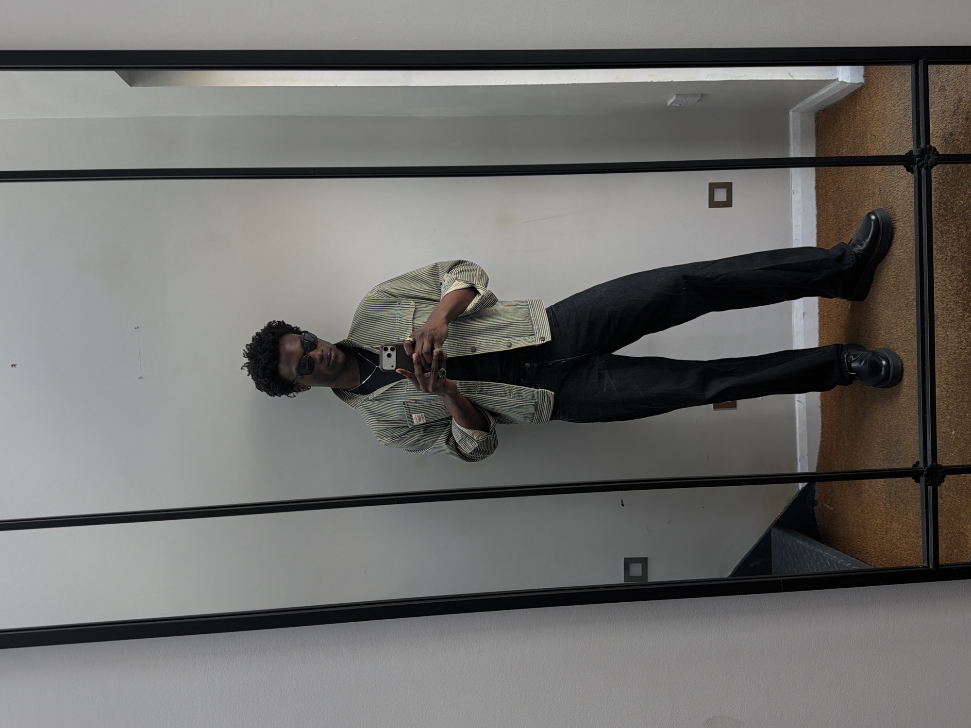 A person, likely male, is captured in a full-length mirror selfie, positioned centrally within the frame which is rotated 90 degrees counter-clockwise. The individual has dark, curly hair and is wearing dark sunglasses, obscuring their eyes and much of their facial expression, which appears neutral.
They are dressed in a casual, workwear-inspired ensemble. This includes a light-colored, possibly green or cream, striped jacket with sleeves cuffed at the wrists. A small, red rectangular label or tag is visible on the jacket's left chest pocket, though its text is illegible. Beneath the jacket, a dark t-shirt is worn. The lower body is clad in dark, straight-leg trousers and black, chunky-soled shoes, possibly platform or combat boots. Several rings are visible on the fingers of the hand holding the smartphone.
The smartphone, with a silver or white back, is held up to take the photograph, covering the person's mouth area.
The background reflected in the mirror consists of plain, light-colored walls, likely off-white or light grey. On the right side of the lower frame, a section of brown carpet is visible on the floor, next to a white wall trim that suggests a corner or doorway. A small, dark square is also present on the wall. The mirror itself is framed in black metal, featuring a decorative, intertwined scroll-like embellishment on the right side.
The setting is an indoor space, providing no specific time of day or weather cues, other than the location context of London, United Kingdom.