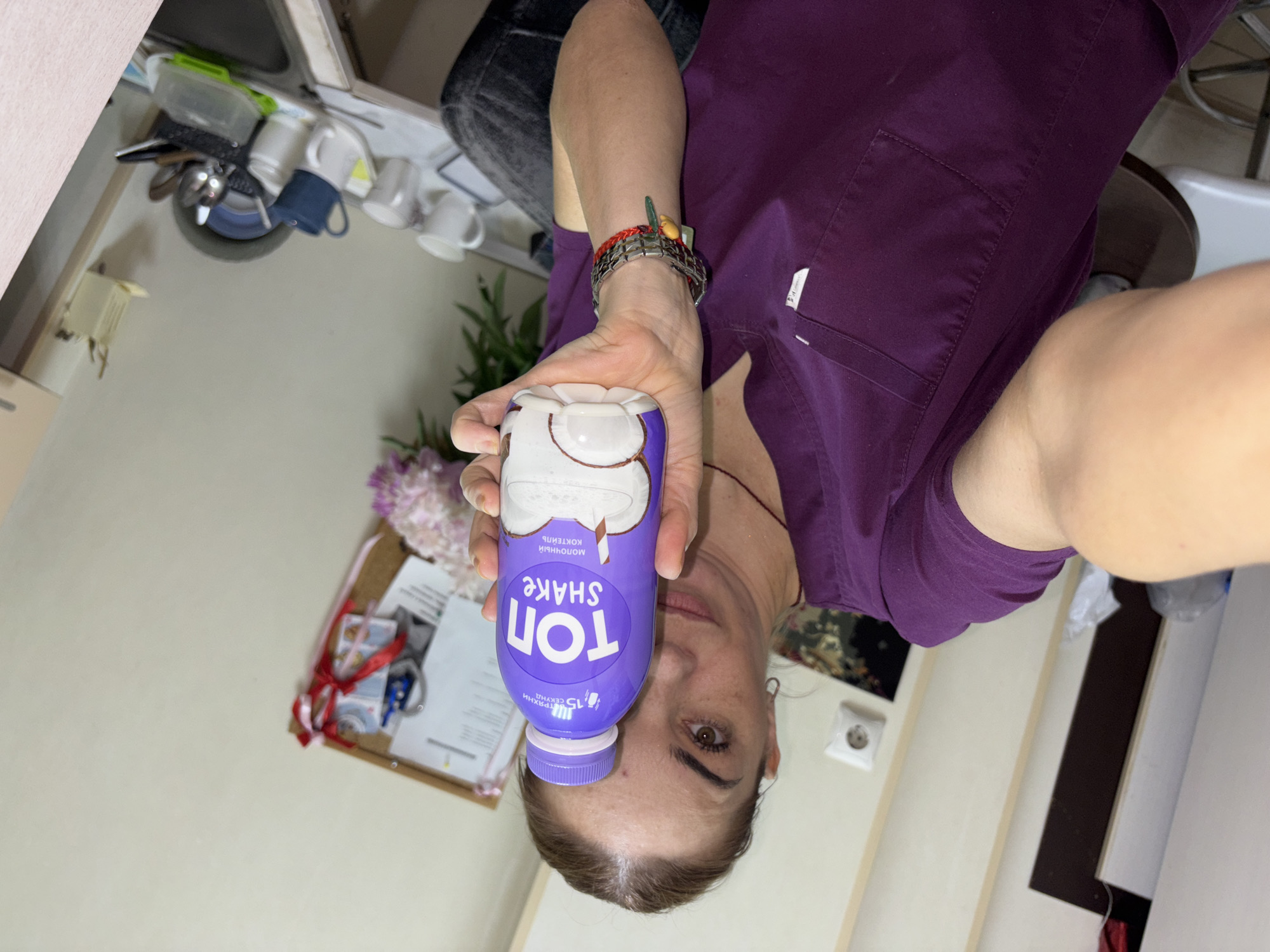 A woman in a purple scrub top is holding up a bottle of "TOP SHAKE" coconut milk. She is in a room with a corkboard on the wall with papers and ribbons attached. There are also mugs, plates, and cutlery on a counter in the background. The location is Medovoye, Russia.