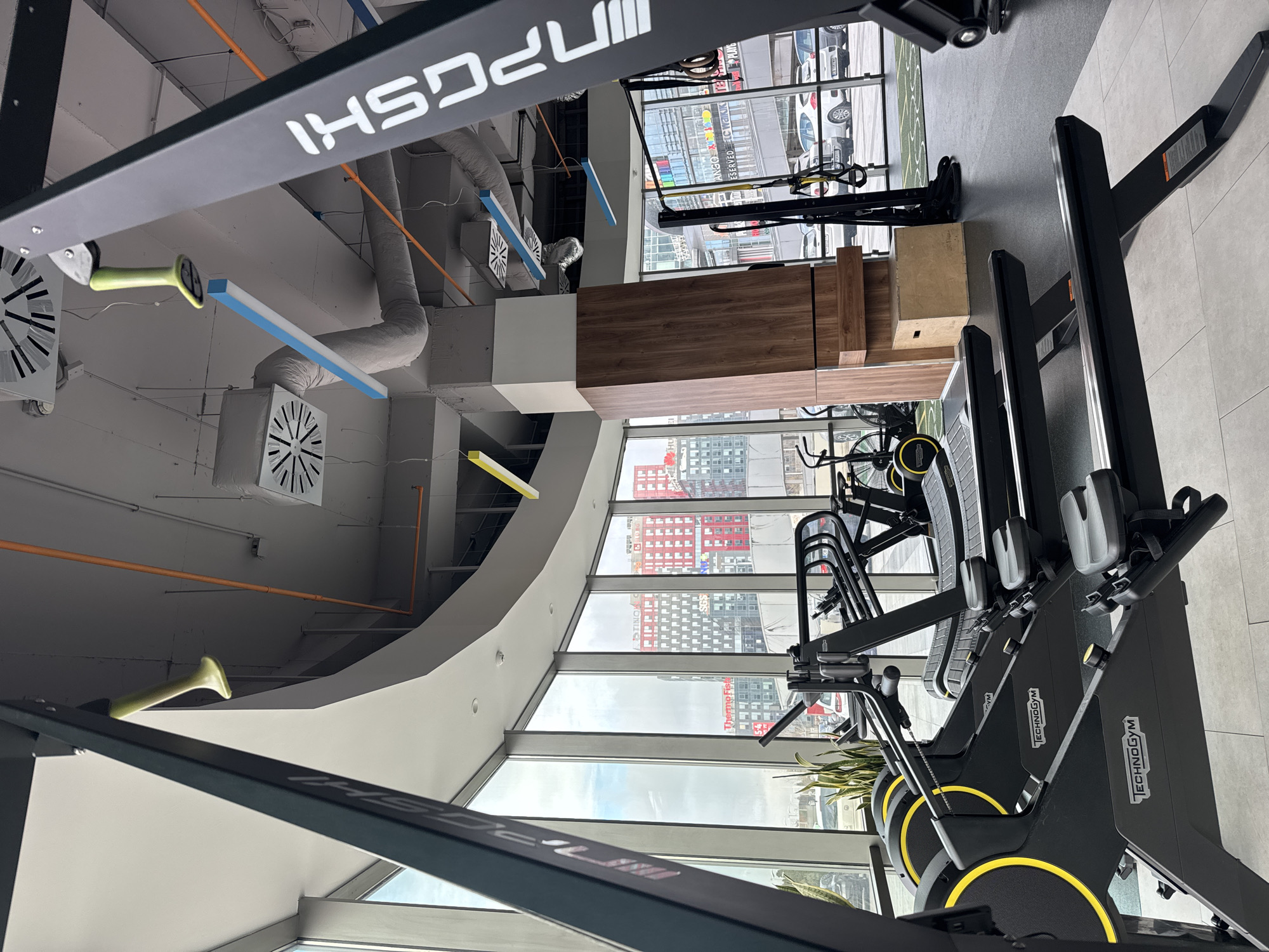 This indoor shot depicts a gym in Sofia, Bulgaria. The camera is oriented to show a scene dominated by indoor fitness equipment, specifically, "Technogym" treadmills, arranged in a row against a backdrop of large windows that reveal an urban landscape. Visible through the windows are buildings with distinctive red and grey facades, typical of Sofia. The foreground is dominated by the black metal framing of the gym equipment. In the background, beyond the treadmills and windows, the cityscape emerges, including roads and parked vehicles. The ceiling of the gym is visible, featuring ventilation ducts and colorful linear lighting in orange, blue and yellow.