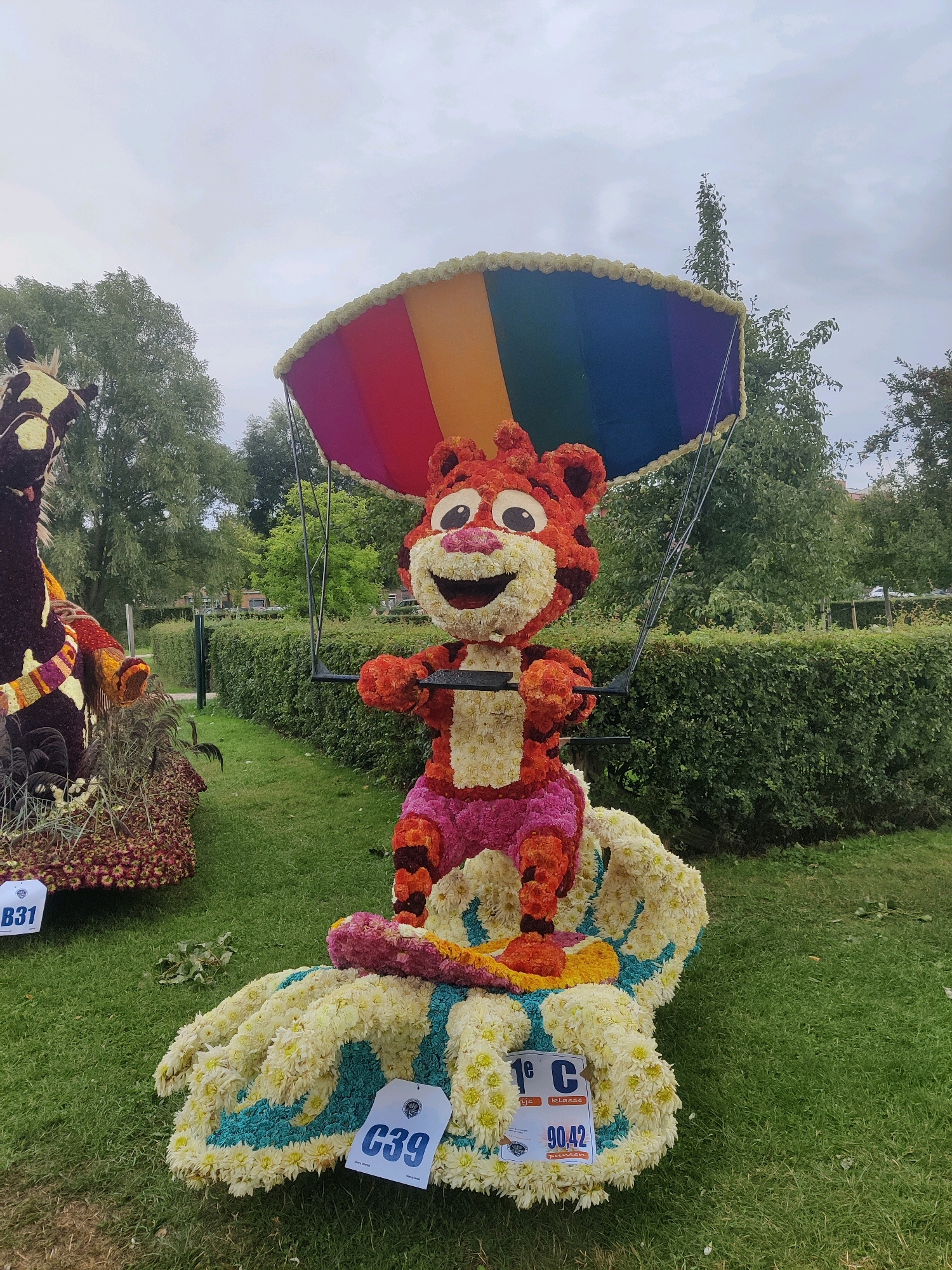 The image depicts a large floral float shaped like a cartoon tiger wearing shorts, suspended by wires from a colorful parachute made of flowers. The tiger is positioned on top of a floral sculpture resembling a giant hand. Two blue tags are visible on the ground in front of the hand sculpture, one labeled "C39" and another with "1e C" and "90,42". To the left, another floral sculpture of an animal, possibly a giraffe, is partially visible. The float is situated outdoors on a grassy lawn, with a neatly trimmed hedge and trees in the background under a cloudy sky. The overall impression is of an outdoor exhibition or parade, likely part of a floral festival. The location is identified as Winterswijk, Netherlands.