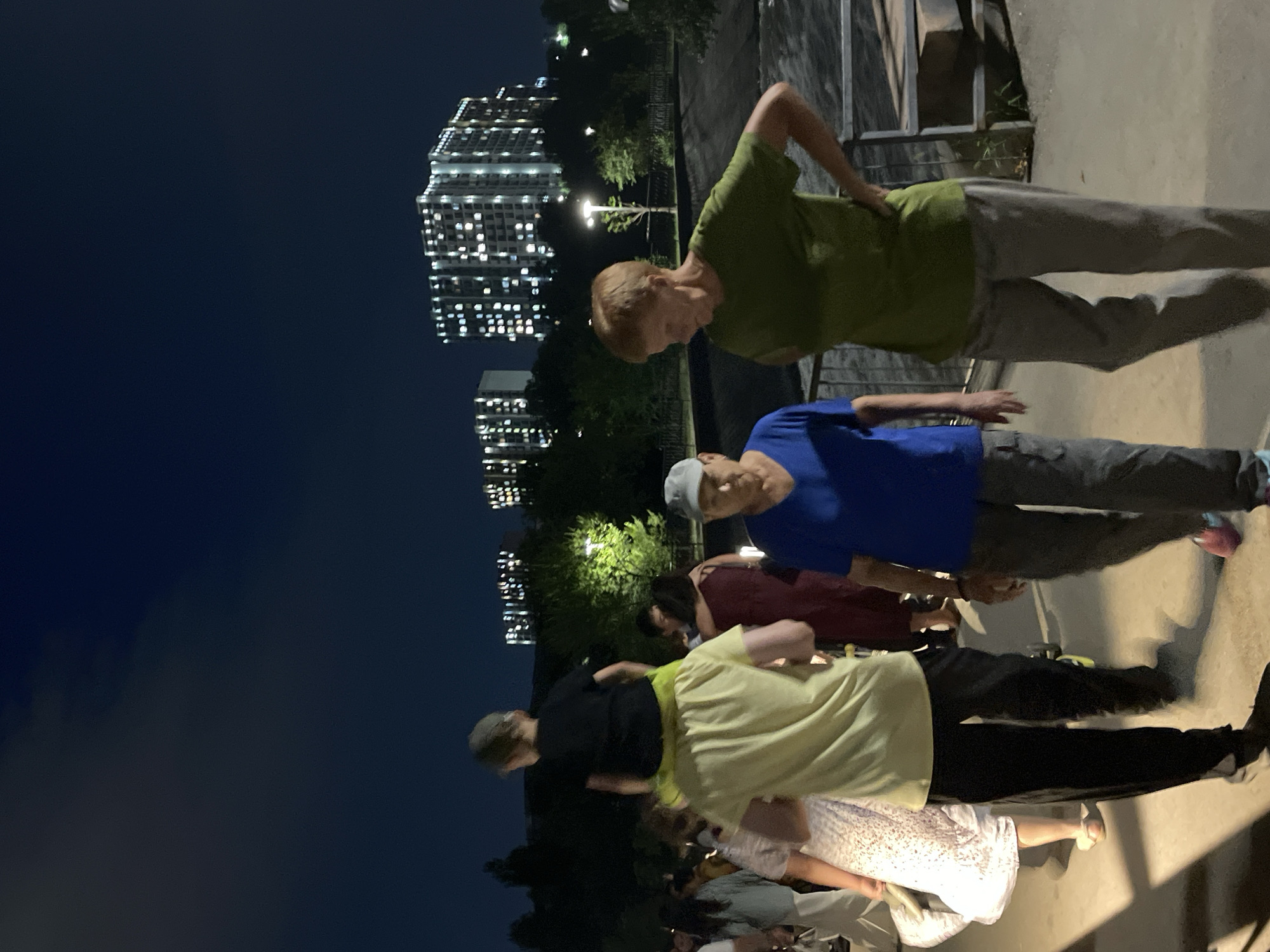 The image depicts an outdoor scene at dusk or night, with several people walking on a pathway. In the foreground, two older individuals are visible, one wearing a blue t-shirt and a cap, and another in a green t-shirt and light-colored pants. Behind them, a child is being carried on the shoulders of an adult wearing a yellow shirt and black pants. Other individuals and parts of their clothing are visible further back in the crowd. The background features silhouetted trees and illuminated multi-story buildings with many lit windows, suggesting an urban environment. The overall impression is of people gathering or moving in a public space after dark, with city buildings as a backdrop. The mention of Almaty, Kazakhstan, provides a specific geographical context for the urban setting.