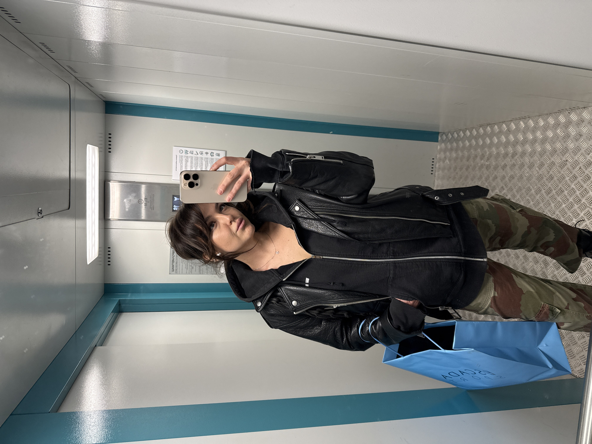The image displays a full-body mirror selfie of a woman inside an elevator. The woman, with dark hair and bangs, is seen looking towards her light-colored smartphone, which has a triple camera setup and is held in her right hand to take the picture. She is dressed in a black leather jacket worn over a black hooded top and camouflage-print cargo pants. A delicate necklace with a small pendant adorns her neck. In her left hand, she holds a light blue paper shopping bag, bearing the mirrored text "ESCADAG," which is likely the brand "ESCADA."
The elevator's interior features white or light grey walls with a prominent blue-green horizontal stripe running along the mid-section. The floor is covered with a silver diamond plate pattern. Various elements of the elevator are reflected in the mirror, including a vertical fluorescent light fixture, a control panel with multiple buttons, and a small sign displaying several icons and some mirrored Cyrillic text, which is likely related to elevator operations or safety instructions. The overall setting appears clean and functional. The woman's expression is neutral, indicative of posing for a photograph. The location context provided for this image is Krasnoyarsk, Russia.