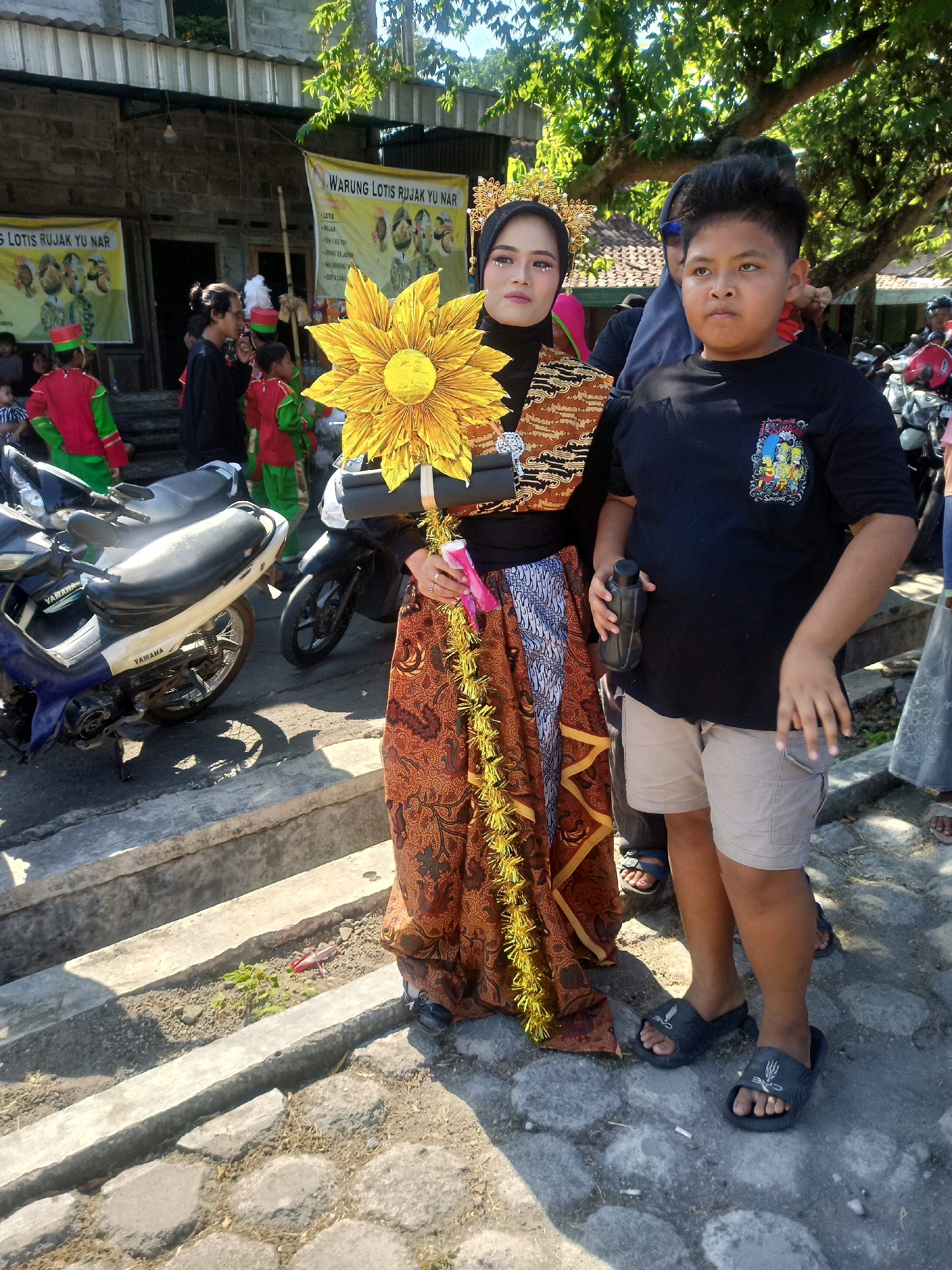 The image captures a street scene in Sumberlawang, Indonesia, under bright daylight. Two individuals are prominently featured in the foreground: a young woman adorned in traditional attire and a young boy. The woman, wearing a black hijab and a golden headdress, holds a large, golden sunflower prop. She is dressed in a patterned brown and orange skirt and a black top, with a decorative sash across her chest. To her right, the boy wears a black t-shirt with a "The Simpsons" graphic and light brown shorts. He is holding a dark water bottle. Behind them, several other people are visible, including children in red and green uniforms and adults. Two motorcycles are parked in the middle ground. In the background, a building with signage that reads "WARUNG LOTIS RIJAL YU NAR" is visible, suggesting a food stall or shop. The environment consists of a paved ground, concrete steps, and trees providing shade. The overall atmosphere appears to be that of a local event or gathering.