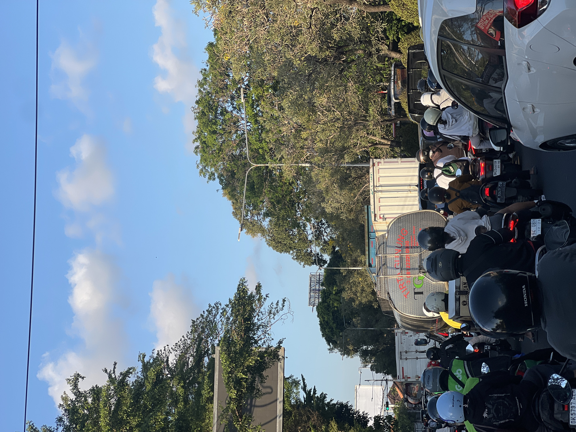 The image depicts a traffic jam in Denpasar, Indonesia, with numerous motorcycles and cars on a road lined with lush green trees. The sky is a clear blue with scattered white clouds. In the foreground, the back of a white car is visible, partially obscuring the view of the vehicles behind it. A line of motorcycles stretches into the distance, with many riders wearing helmets of various colors, including white, black, and green. Some motorcycles have passengers. A large, silver tanker truck with red lettering that reads "TIRTA GUNUNG TG" is positioned in the middle ground, alongside white container trucks. The overall scene suggests a typical urban environment with heavy traffic and abundant vegetation.