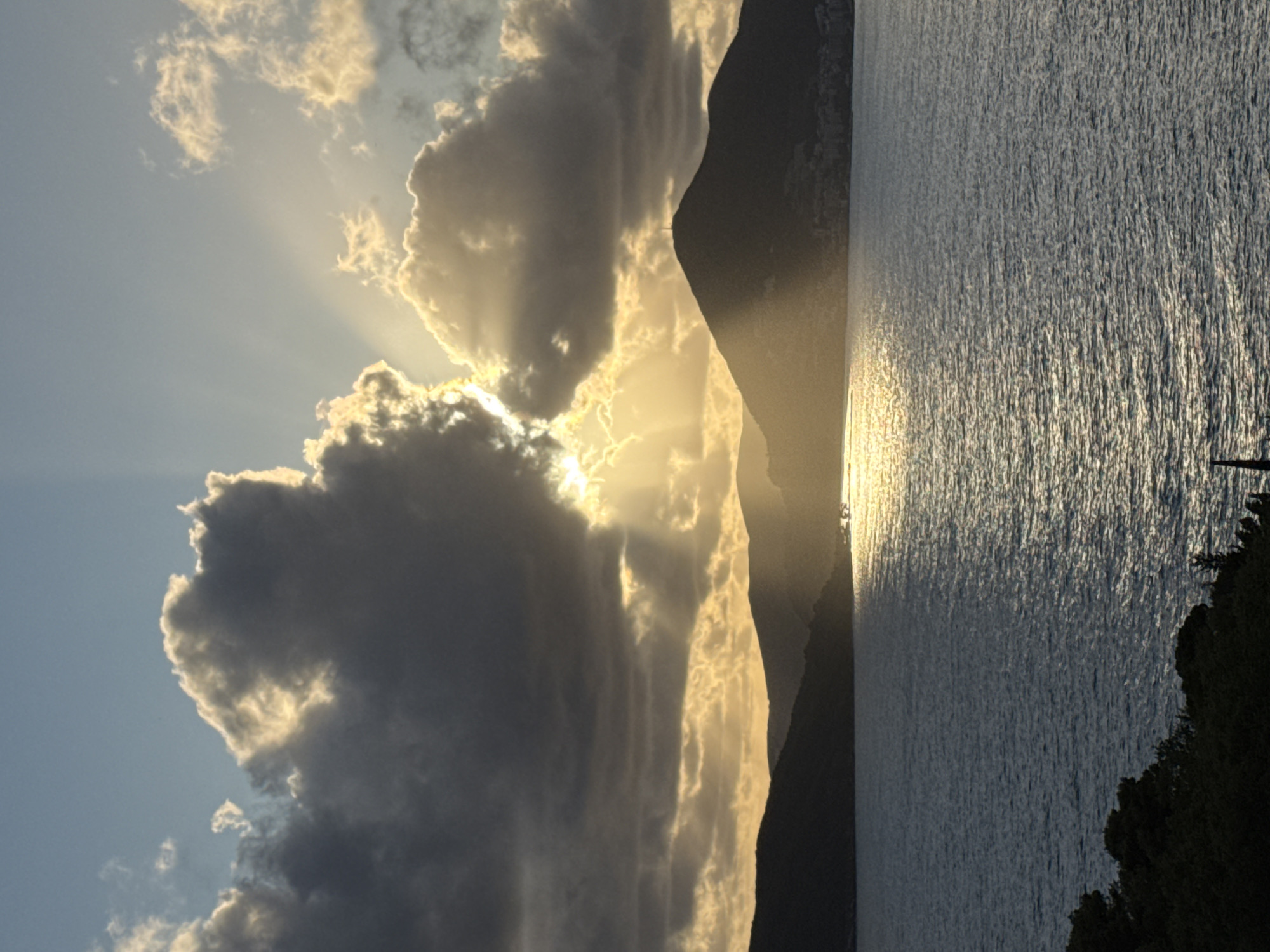 The image shows a sunrise over the sea, likely near Sveti Stefan, Montenegro. Light rays stream through clouds to illuminate the water. The sun's reflection creates a bright line on the sea's surface, with darker mountains on the left and foliage on the right. No people are visible.