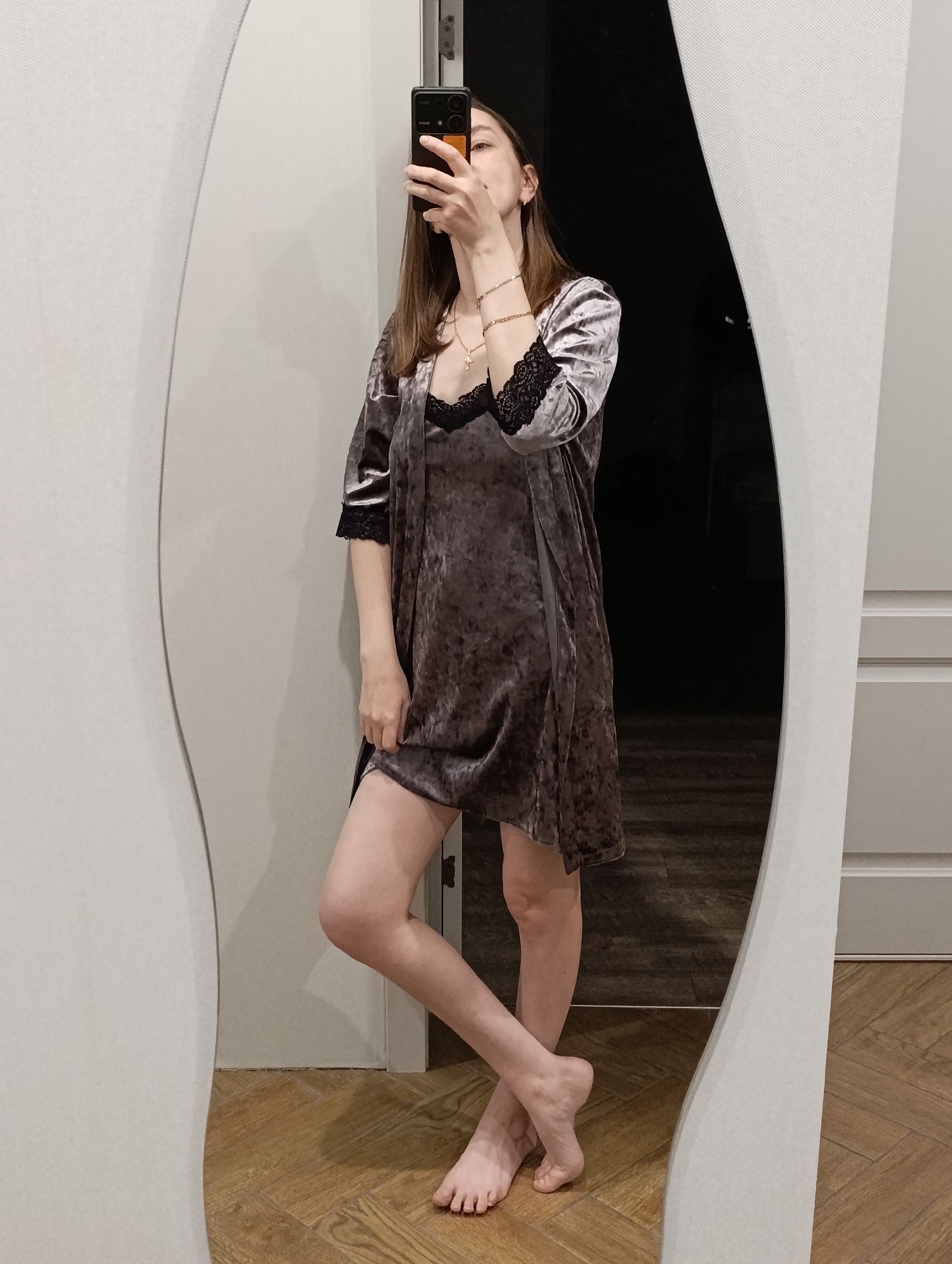 A full-length mirror selfie captures a young woman standing indoors in Chaikovsky, Russia. She holds a black smartphone with an orange accent, obscuring most of her face, as she takes the picture in a mirror with a distinctive curved white frame. The woman has long brown hair and is dressed in a two-piece velvet pajama set in a muted purple-gray hue, adorned with black lace trim on the sleeves of the robe and the neckline of the nightgown. She is barefoot, with one leg slightly bent and crossed at the ankle. Accessories include a delicate gold necklace with a small cross pendant and a gold bracelet on her left wrist. The flooring is light-colored wooden parquet or laminate. In the reflection, the background behind the woman appears dark, suggesting an unlit room or hallway, while the wall where the mirror is positioned is a plain light gray or white. The lighting is consistent with an indoor setting. No visible text is present in the image.