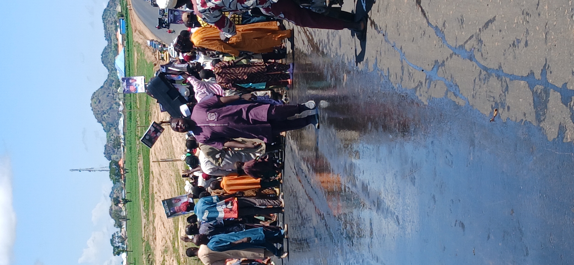 The image depicts a street scene in Kayarda, Nigeria, on a sunny day with a clear blue sky. The foreground shows a wet asphalt road, reflecting the sky and the people. A diverse group of people, predominantly men, are gathered on the road and the adjacent sandy shoulder. They are dressed in various traditional African attire, including kaftans, agbadas, and wrappers. Some individuals are holding up signs or posters, suggesting a gathering or procession. In the background, a road stretches into the distance, with vehicles and a telecommunications tower visible. The landscape features rolling hills and scattered buildings, characteristic of a rural or semi-urban setting in Nigeria.