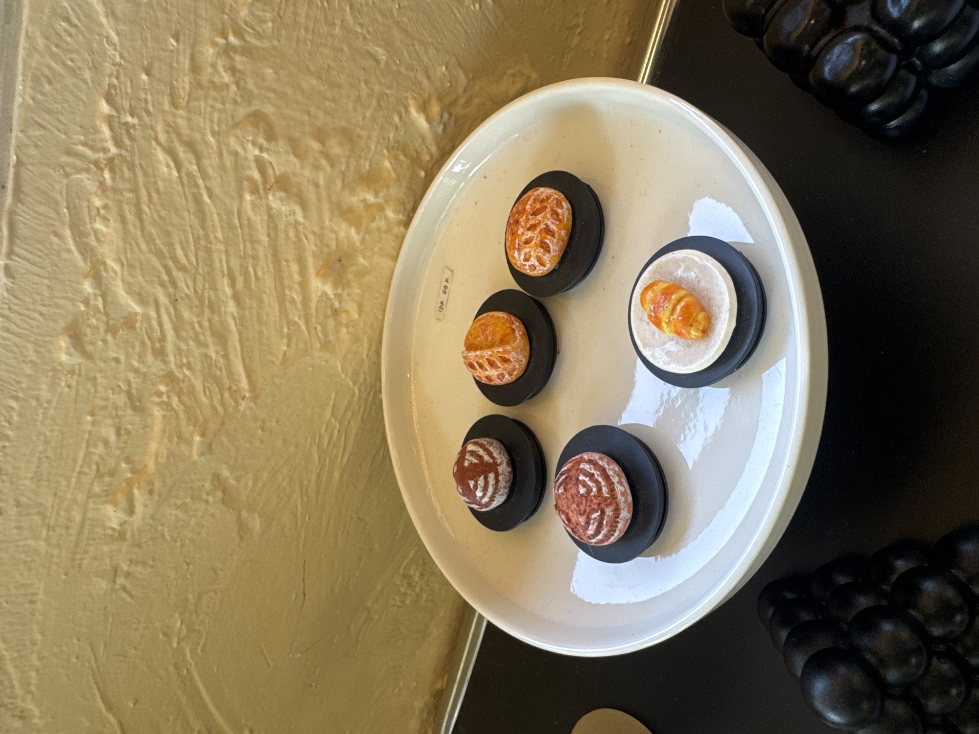 A high-angle shot reveals a beige textured wall adjacent to a white plate holding five small, decorated circular desserts on black pedestals. One dessert is croissant-shaped. Adjacent to the plate, a black surface holds a cluster of shiny, black, spherical objects. The image was taken in Kuta Selatan, Indonesia.