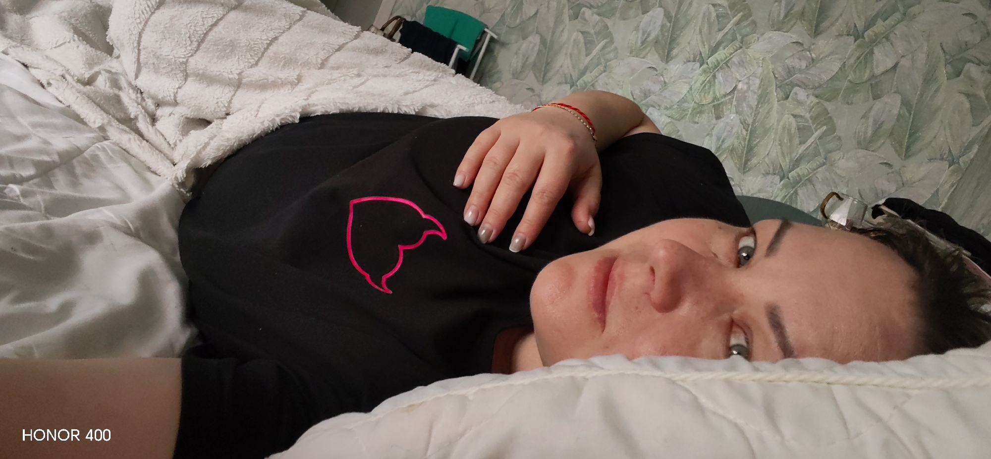 A person is lying in bed under blankets, with a black shirt with a pink logo visible. The background includes a wall with a plant leaf pattern. There is no evidence to suggest this image was taken in the city of Noviy, Russia. The phone brand "HONOR 400" is shown on the bottom left corner.