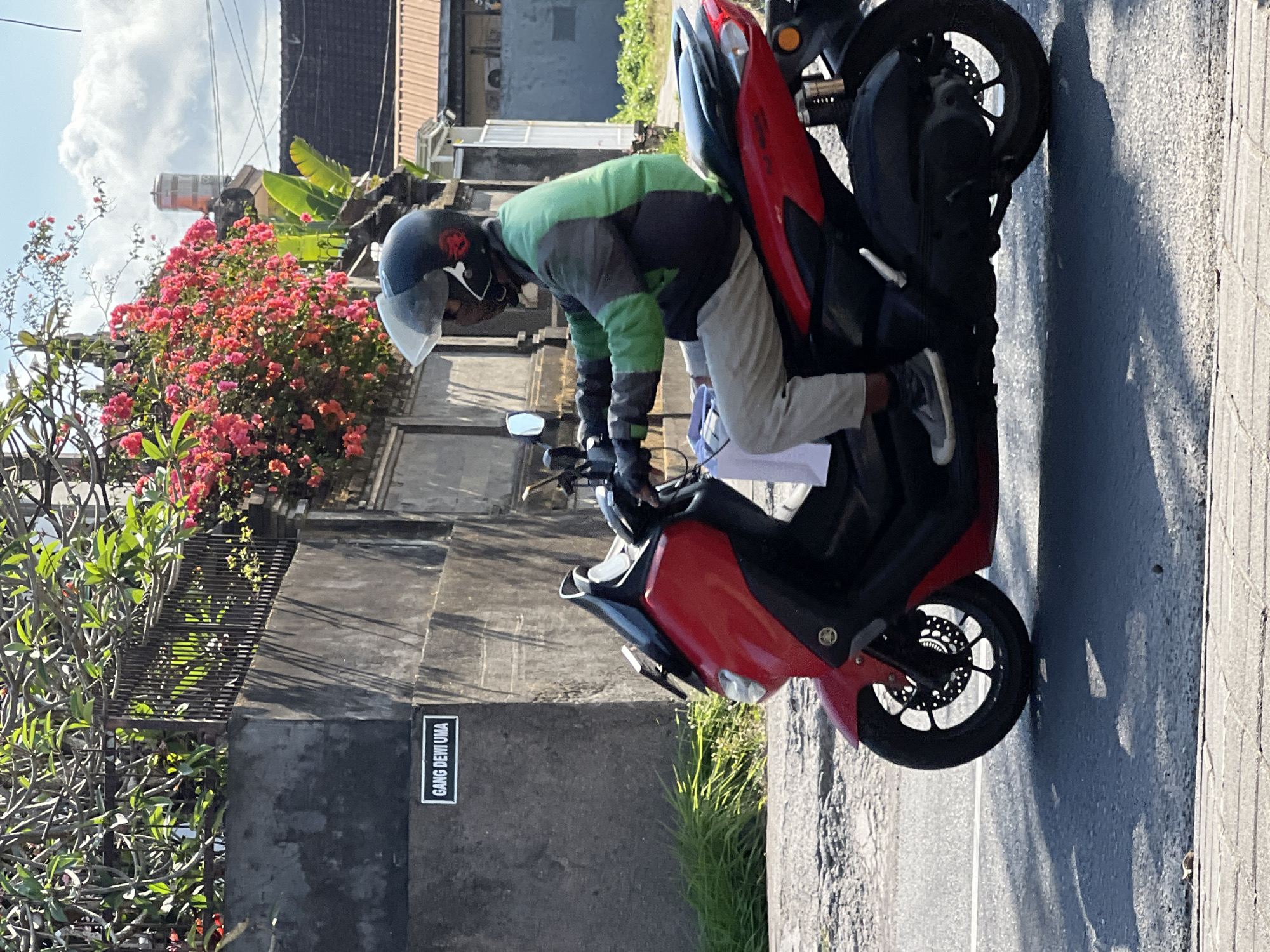 A person wearing a helmet and a green and gray jacket rides a red scooter on a paved road in Denpasar, Indonesia. The scooter has a black seat and a black windshield. The person is wearing gray pants and dark shoes. In the background, there is a gray wall with a sign that reads "GANG DEWI UMA". There are also trees with pink flowers and green leaves growing on top of the wall. The sky is blue with some clouds. The sun is casting shadows on the road.