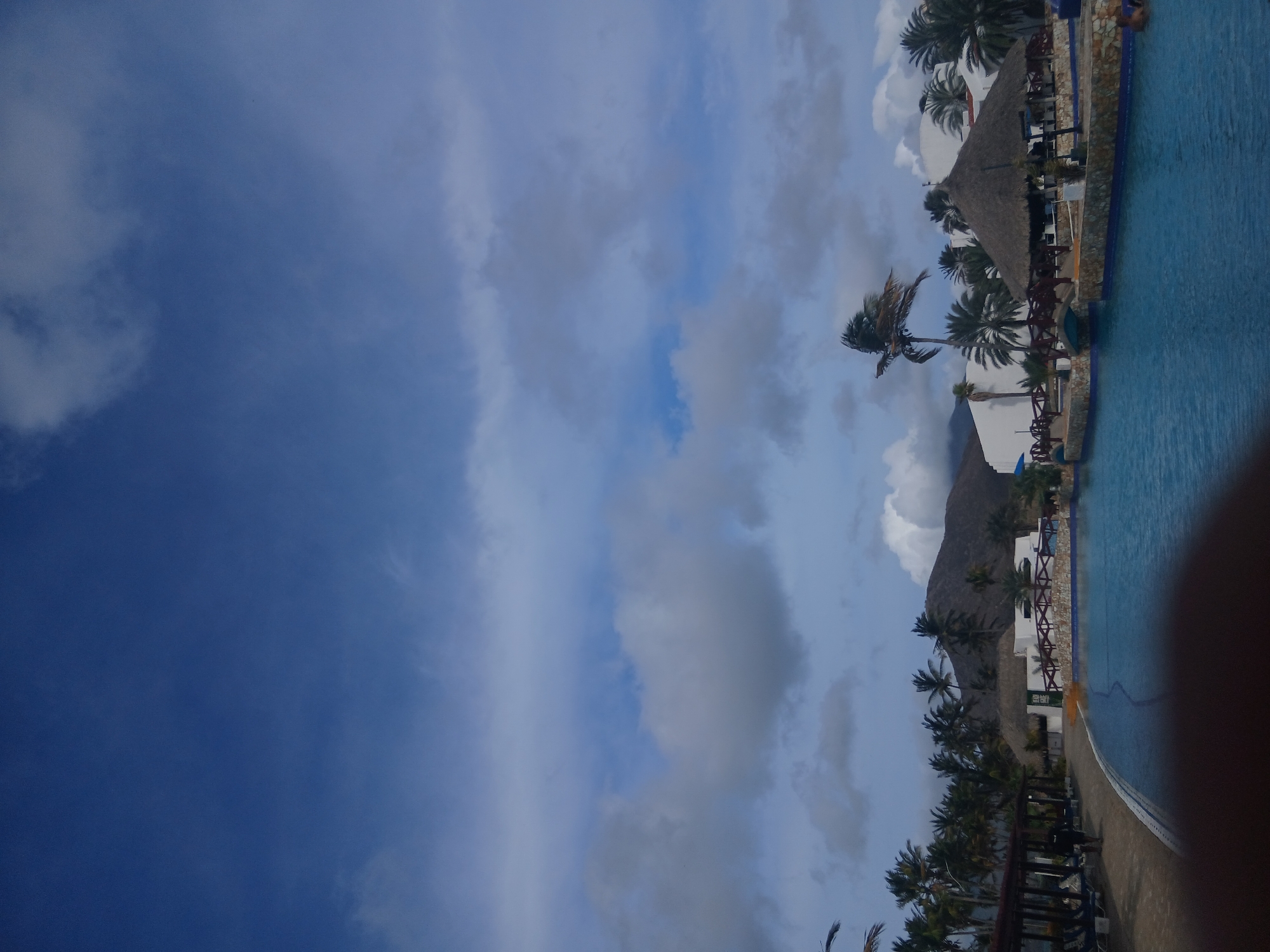 The image shows a swimming pool area in Altagracia, Venezuela. The pool is turquoise blue and runs along the right side of the frame. Palm trees line the side of the pool, with some thatched-roof structures providing shade. White buildings are visible behind the palm trees. In the background, a mountain rises, partly obscured by clouds. The sky is a mix of blue and white clouds.
