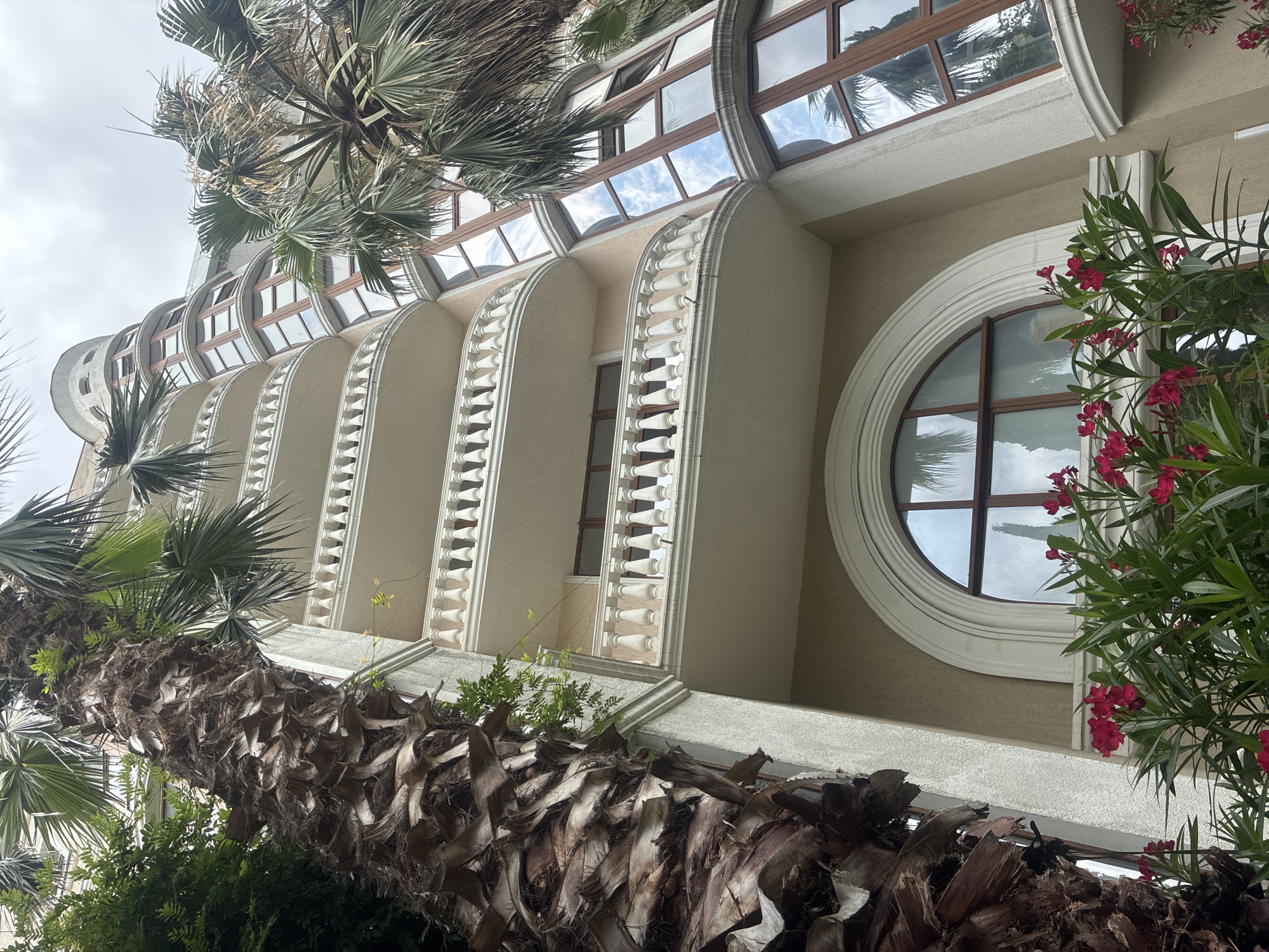 The image captures a low-angle view of a modern, multi-story building with curved balconies, situated in Sochi, Russia. The building's facade is a light beige color, contrasting with white balustrades and a large, arched window with dark wooden trim. The windows on the upper floors are also framed in dark wood and are set into curved sections of the building, reflecting the cloudy sky and palm trees. In the foreground, a large, weathered palm tree trunk is prominently featured on the left, with lush green foliage and vibrant pink flowers growing at the base of the building. The overall impression is one of a luxurious resort or hotel, typical of the subtropical climate of Sochi. There are no people visible in the image, and the scene appears to be static and undisturbed. The lighting suggests it is daytime, with overcast conditions.