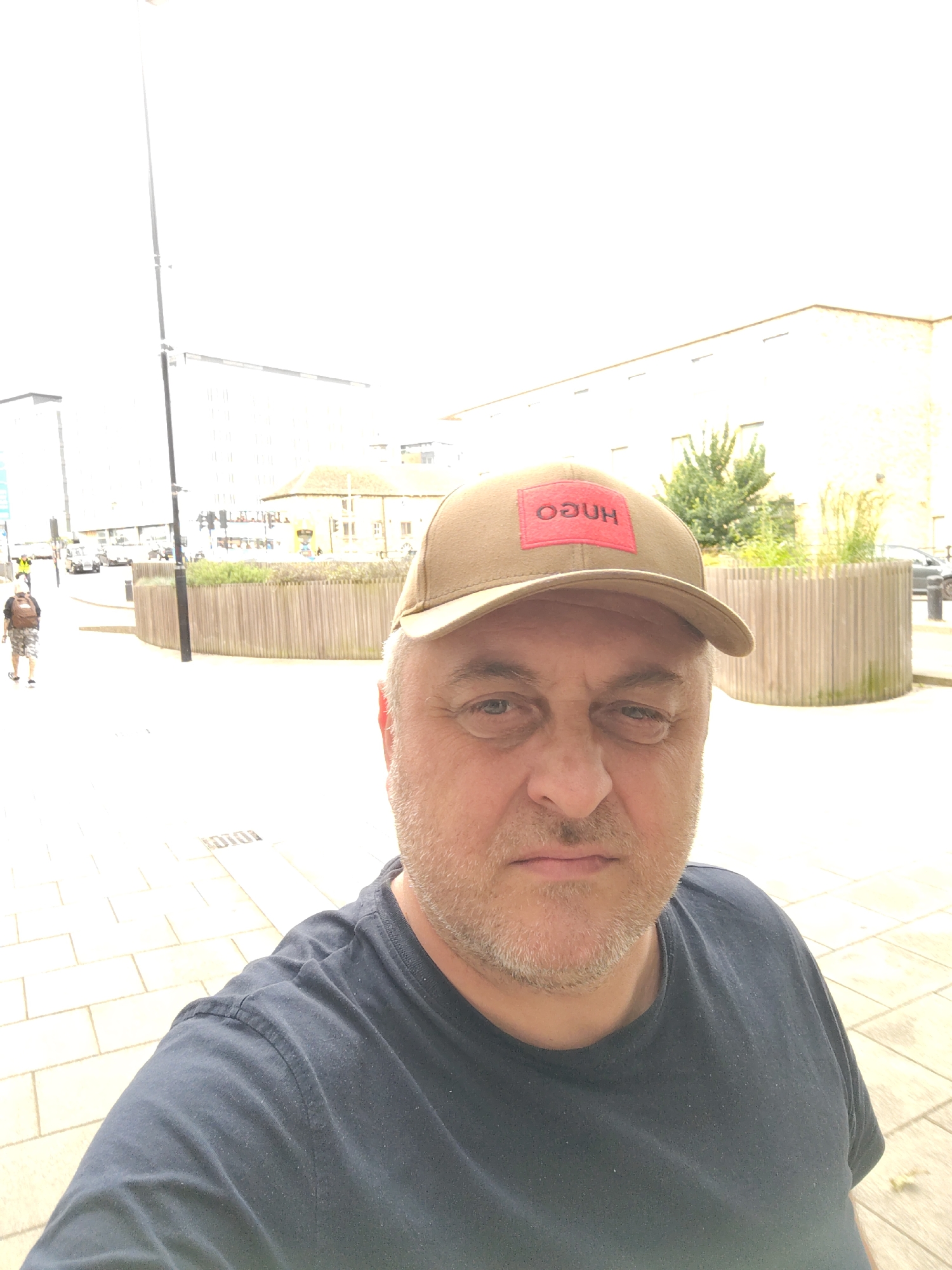 The scene depicts an outdoor urban environment in Peterborough, United Kingdom, during daylight hours. In the immediate foreground, a middle-aged male subject is positioned, facing the camera directly. He is Caucasian, with fair skin, short grey hair visible at the sides, and light grey stubble or a short beard. He wears a dark navy blue crew-neck t-shirt and a beige baseball cap. The cap features a red rectangular patch on the front with the white word "HUGO" inscribed, appearing upside down from the viewer's perspective. The subject's expression is neutral, with slightly narrowed eyes, suggesting a direct gaze into the camera.
The background reveals a paved pedestrian area transitioning into a street. To the far left, a tall, slender black lamppost extends vertically. Beyond the lamppost and to the left, modern, light-colored multi-story buildings are visible under bright, slightly overexposed skies. A person wearing a backpack and shorts is seen walking away from the camera on the paved surface, to the left of the main lamppost. Further down the street, multiple vehicles, including a small white utility vehicle, are present.
Centrally and extending to the right, a circular planter with vertical wooden slat fencing encloses green vegetation. Behind this planter, a light-colored building with a more traditional architectural style, possibly stone or light brick, features several recessed rectangular openings or windows along its upper facade. Additional green foliage and distant structures are visible behind this building. The ground consists of light-colored, rectangular paving stones, with faint markings embedded in the surface, such as "10101" visible on the lower left. The overall lighting is bright, indicating an outdoor setting, potentially under an overcast or bright, hazy sky, causing some highlights to be blown out.
