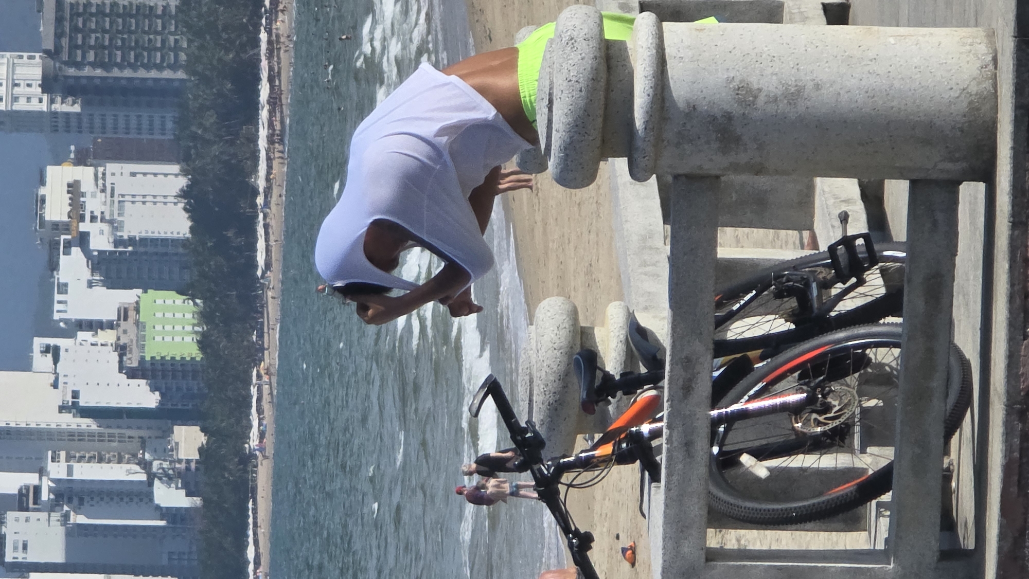 The image depicts a beach scene in Nha Trang Ward, Vietnam. A person wearing a white shirt and bright green shorts stands on a concrete structure facing the ocean. The person is bent over, possibly rinsing off or interacting with the water. A bicycle is parked nearby, leaning against the concrete structure. The beach and ocean extend into the background, with buildings and vegetation lining the shoreline.