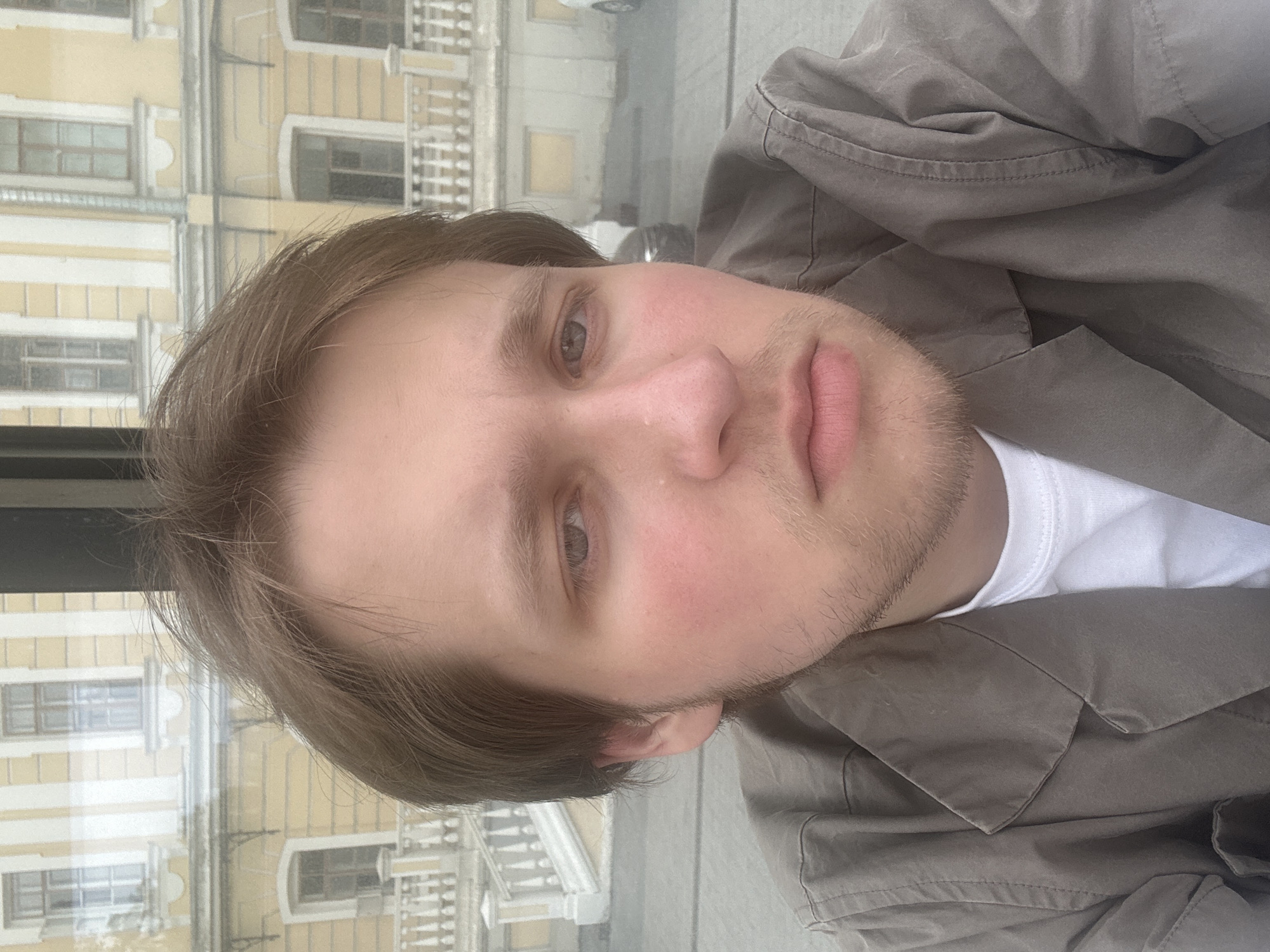 A selfie taken by a fair-skinned person with short brown hair and stubble. They are wearing a white t-shirt and a gray-brown jacket. The background shows a building with yellow walls and multiple windows, seemingly reflected in a glass surface. The location appears to be an urban area in Unknown, Unknown.
