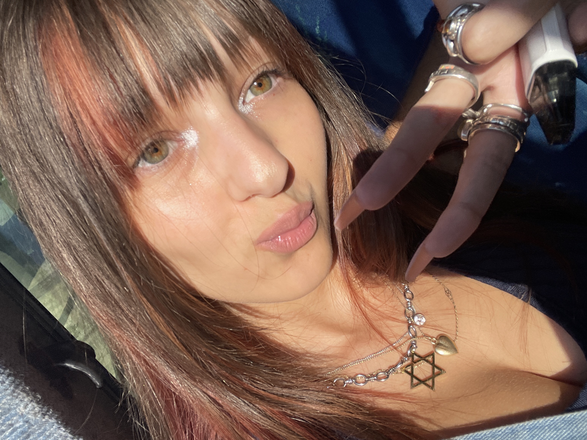 A close-up selfie captures a young woman's face from a high angle, with her body angled downwards. Her right hand, adorned with multiple silver rings, is raised near her face, holding a white vape device. She has long, brown hair with reddish highlights and bangs. Her makeup includes shimmery eyeshadow and lip gloss. She wears a layered necklace with a clear pendant and a Star of David charm. The background is dark and out of focus, suggesting an indoor or dimly lit outdoor environment. The image does not provide specific geographic information to determine its location within Petah Tikva, Israel.