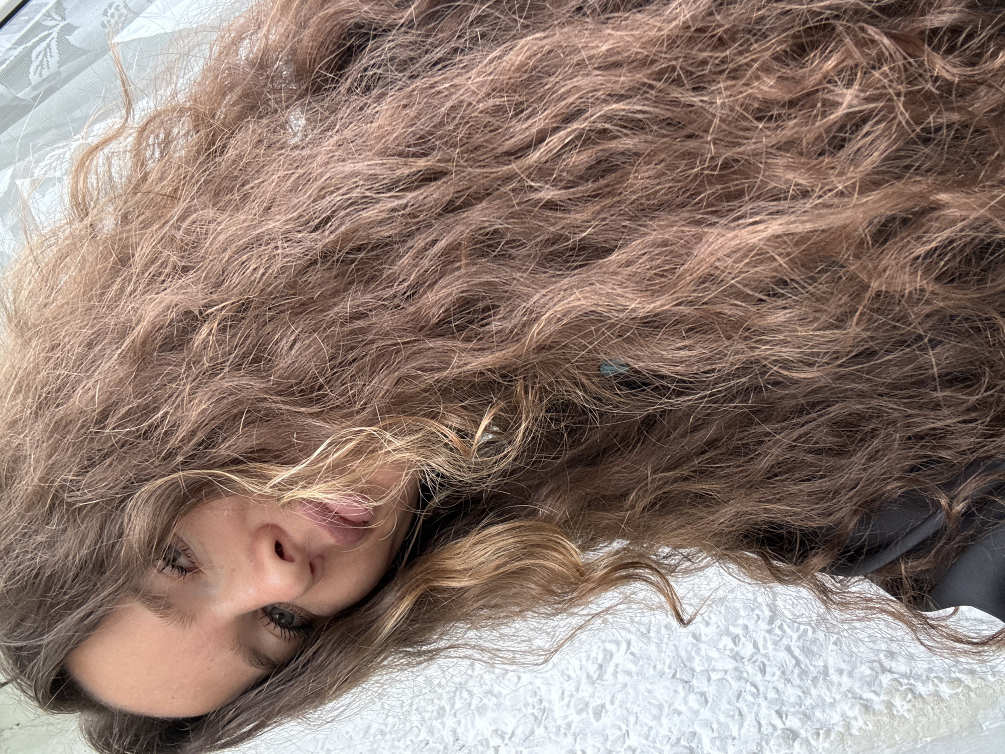 A close-up, low-angle selfie shows a young woman with long, wavy, light brown hair. She is looking at the camera with her head tilted. Her eyes are blue-green and her lips are slightly parted, with her tongue just visible. The background consists of a white textured wall to the left and a lace-patterned curtain to the right. There is no discernible location in the city of Turinsk, Russia.
