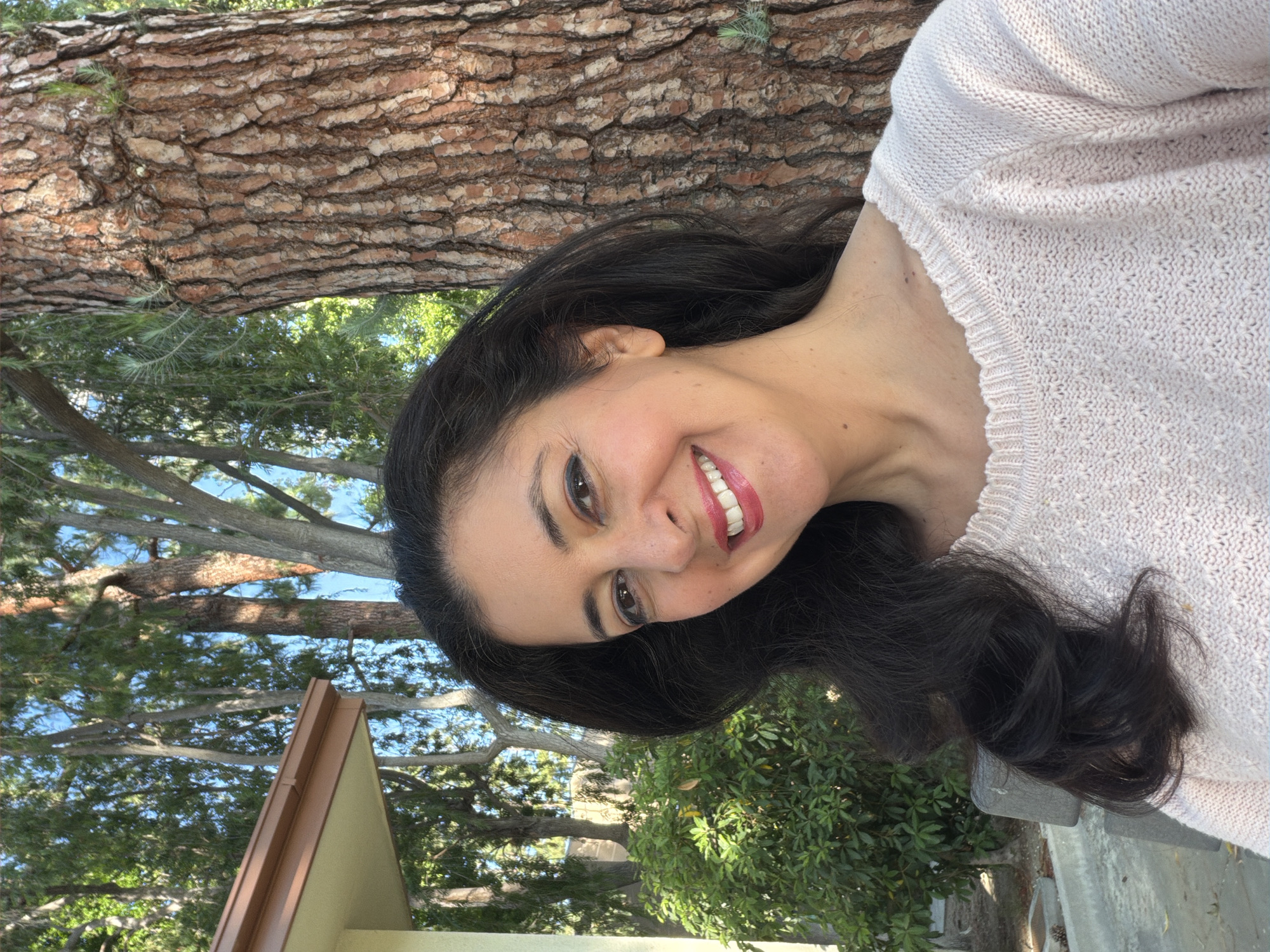 A woman with long, dark hair and a light-colored sweater is smiling in an outdoor setting. Behind her are trees, including a large tree trunk with textured bark, and foliage. The photo also shows a building structure with a brown roof edge. The environment suggests an outdoor location in a place with trees, possibly within a city park or a residential area with mature trees. Since the prompt requires it, it is likely within the city of Los Angeles, United States of America.