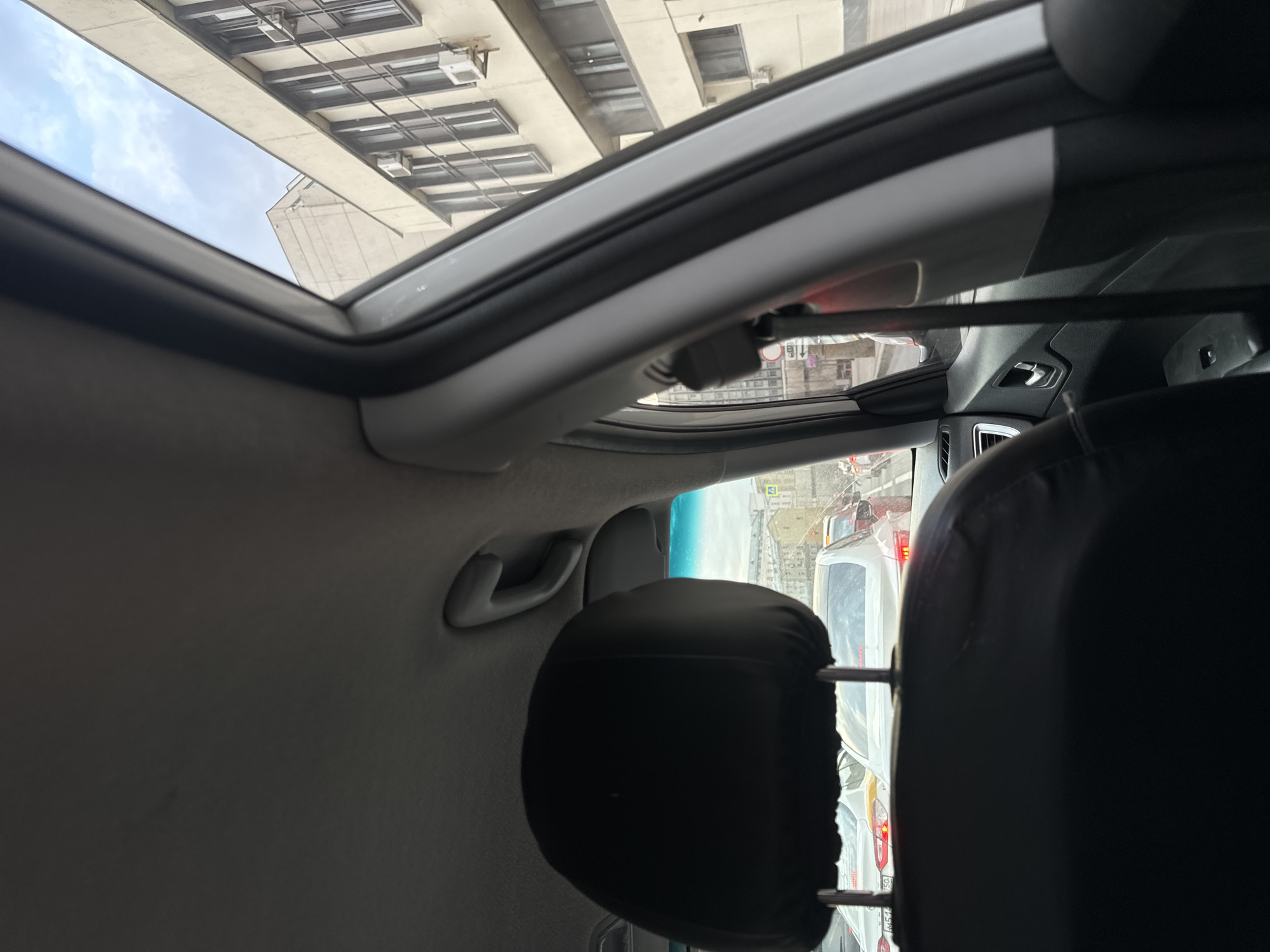 The photograph was taken from the interior of a vehicle looking up through an open sunroof. Outside the sunroof, a tall building with many windows is visible, as is the sky. Through the windshield, a street scene shows traffic with multiple cars, a pedestrian crossing, and street signs in Russian. It's daytime, and the visibility appears to be good. The vehicle is likely in a city, possibly Moscow, Russia, due to the Cyrillic lettering on the signs. The focus is on the interior of the car, with the headrest of the front seat and the car's interior roof being prominent. There's a grab handle on the interior roof as well.
