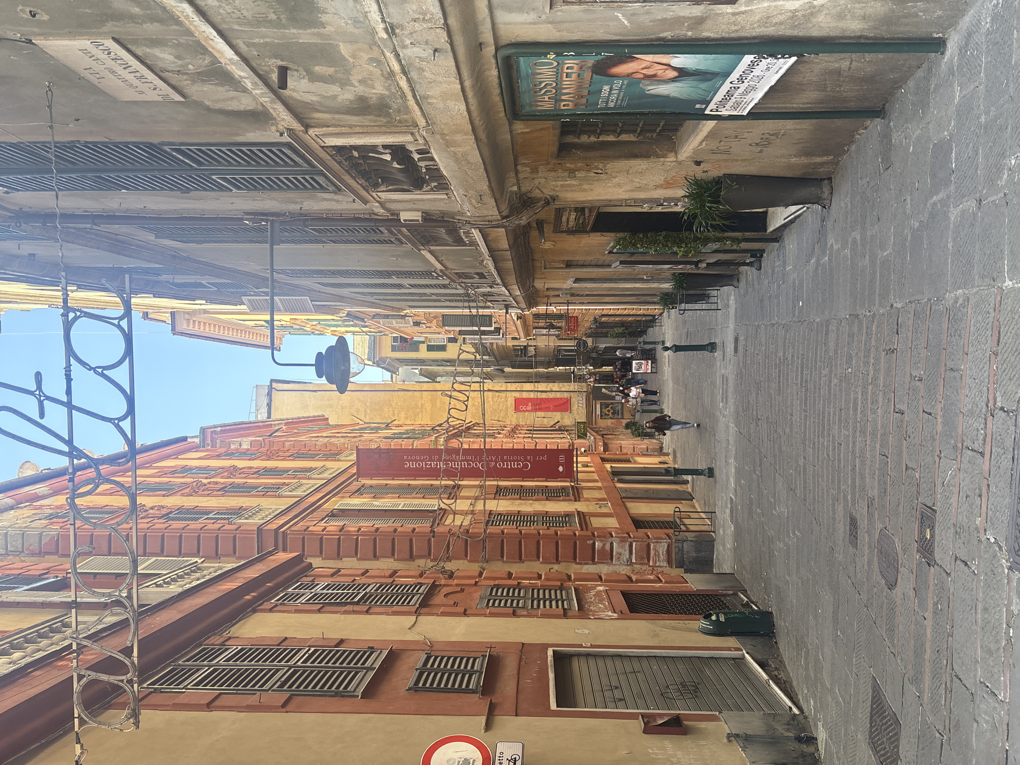 This media file captures a pedestrian street in an old town, likely in Genoa, Italy, given the architectural style and visible Italian text. The street is paved with block-like stones and extends into the distance, flanked by multi-story buildings with weathered facades.
The buildings on the left are primarily in shades of orange, yellow, and red, featuring numerous windows often adorned with closed wooden shutters. A prominent red banner-like sign is affixed to one building, reading "Centro di Documentazione per la Storia l'Arte l'Immagine di Genova," indicating a cultural or historical center. Decorative metal railings are also visible along this side.
On the right side of the street, the buildings appear in more subdued, grayer tones. A noticeable green billboard displays an advertisement for "MASSIMO RANIERI," with the text "TUTTI SOGNI ANCORA IN VOLO E CONTINUANO A VOLARE." Additional text on the billboard specifies a performance at "Politeama Genovese" on "Sabato 1 Maggio 2021 ore 20:30." Below this, a street sign clearly states "VIA AI QUATTRO CANTI DI S. FRANCESCO." Potted plants add greenery along the sidewalk, and faint graffiti, including "NO TAV," is visible on the ground or a wall.
Several individuals can be seen walking casually in the distance, suggesting a normal day with pedestrian activity. The scene is brightly lit by natural daylight, indicating clear and sunny weather. Streetlights are present but unlit. The overall impression is a historic and lively urban thoroughfare.